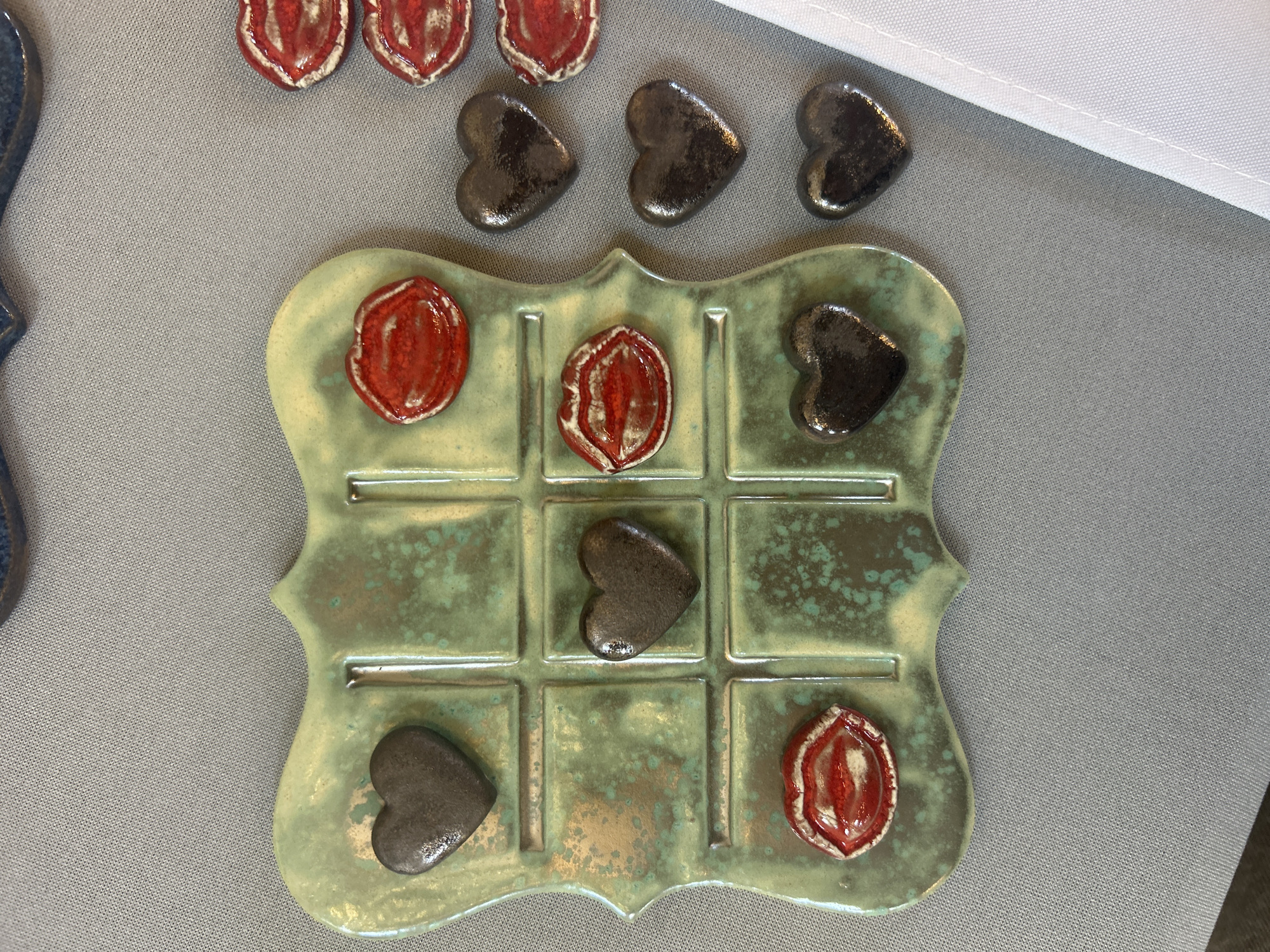 The image shows a ceramic tic-tac-toe board with playing pieces, likely handmade. The board is green with a crackled glaze, and the playing pieces consist of heart-shaped pieces and reddish almond-shaped pieces. A game is in progress on the board. The board is placed on a gray surface. Some playing pieces are lined up next to the game board. A white piece of cloth is on the top right. It is unknown if this image was taken in Sofia, Bulgaria.