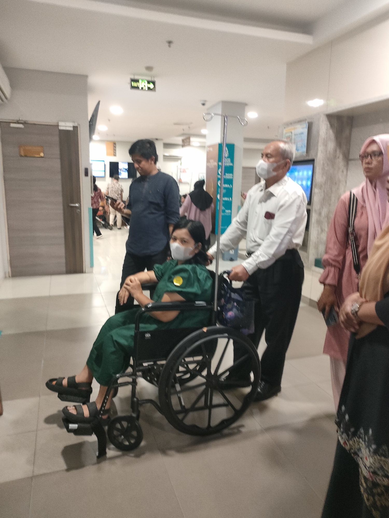 A group of people are in a brightly lit hospital corridor in Pekanbaru, Indonesia. In the foreground, a woman wearing a green uniform and face mask is seated in a wheelchair, her feet resting on the footrests and wearing black sandals. She is being pushed by an older man wearing a white collared shirt and black pants, who is holding the wheelchair handles. Behind them, a man in a dark blue shirt stands looking towards the left. To the right, a woman in a pink hijab and a pink shirt stands holding a black purse, and another person in dark clothing with a patterned skirt is partially visible. In the background, a green "EXIT" sign with an arrow is illuminated on the ceiling. There are doors, medical equipment, and screens visible along the corridor walls.