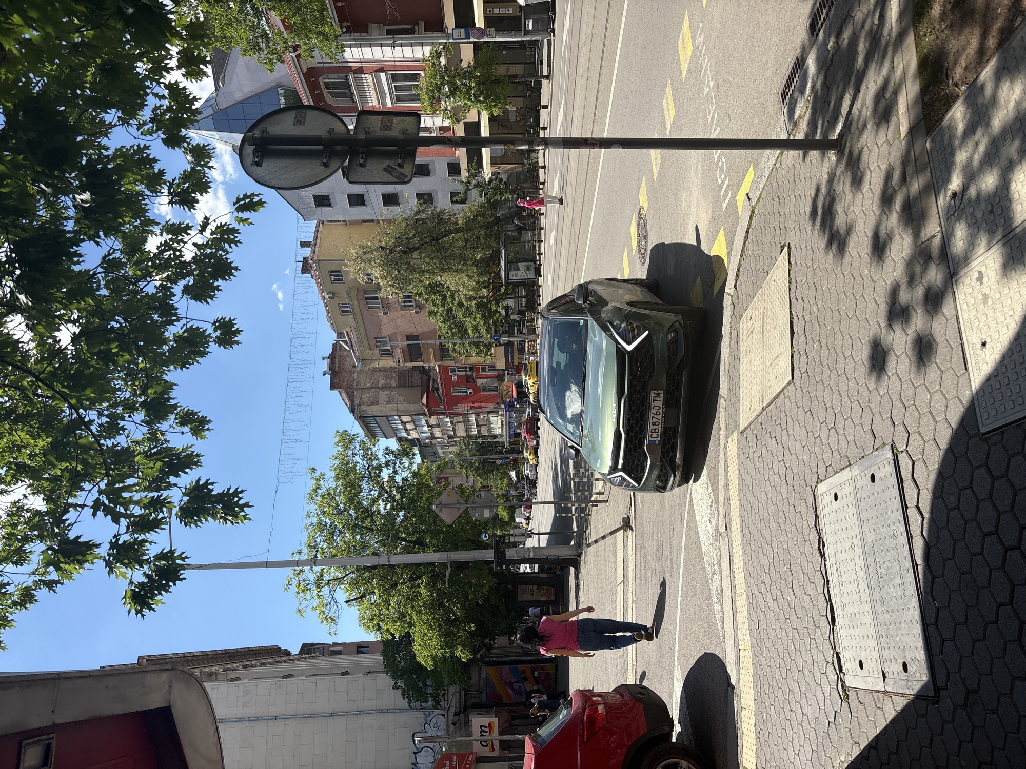 The image depicts a street scene in Sofia, Bulgaria. A pedestrian in a pink shirt and jeans walks on the crosswalk. A dark green car is parked partially on the crosswalk. Buildings line the street, with trees providing shade. A traffic sign is visible. Other cars are parked along the street, and some pedestrians are visible further down the street. The street has marked crosswalks and yellow markers. A red car is parked behind the pedestrian.