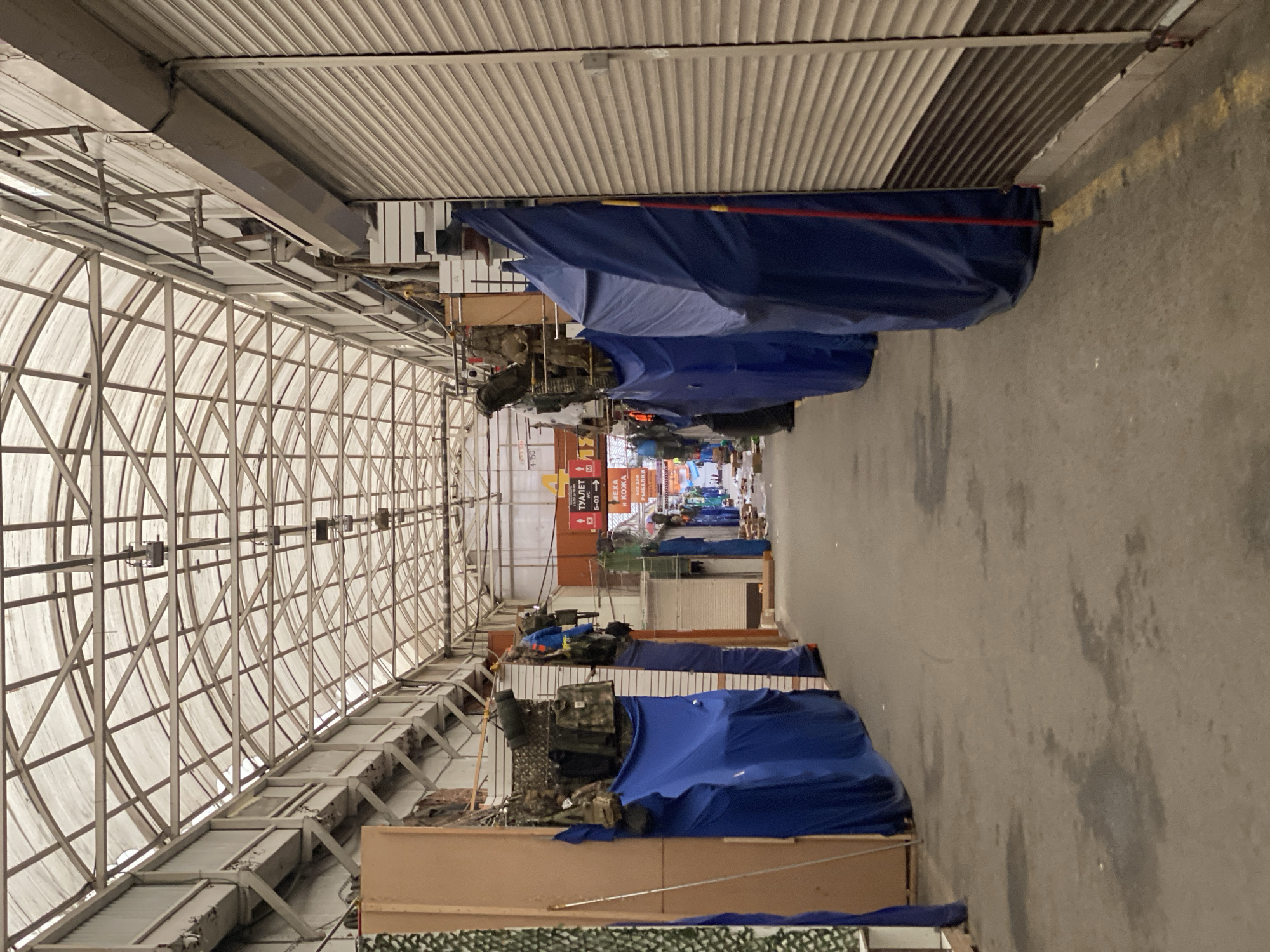 The image depicts an indoor market aisle in Moscow, Russia, likely during off-hours or a slow period. The aisle is covered by a translucent, arched roof supported by a metal frame. On the right side, a row of closed metal shutters lines the market stalls. On the left, market stalls are partially covered with blue tarpaulins, with some goods, including what appears to be military or outdoor equipment, displayed above. Signs in Russian are visible in the background, one of which indicates "ТУАЛЕТ" (Toilet) and another mentions "РЫБАЛКИ" (Fishing). The ground is paved with asphalt and shows some water stains. No people are actively engaged in commerce or visible activity within the aisle, suggesting a quiet or closed state of the market.