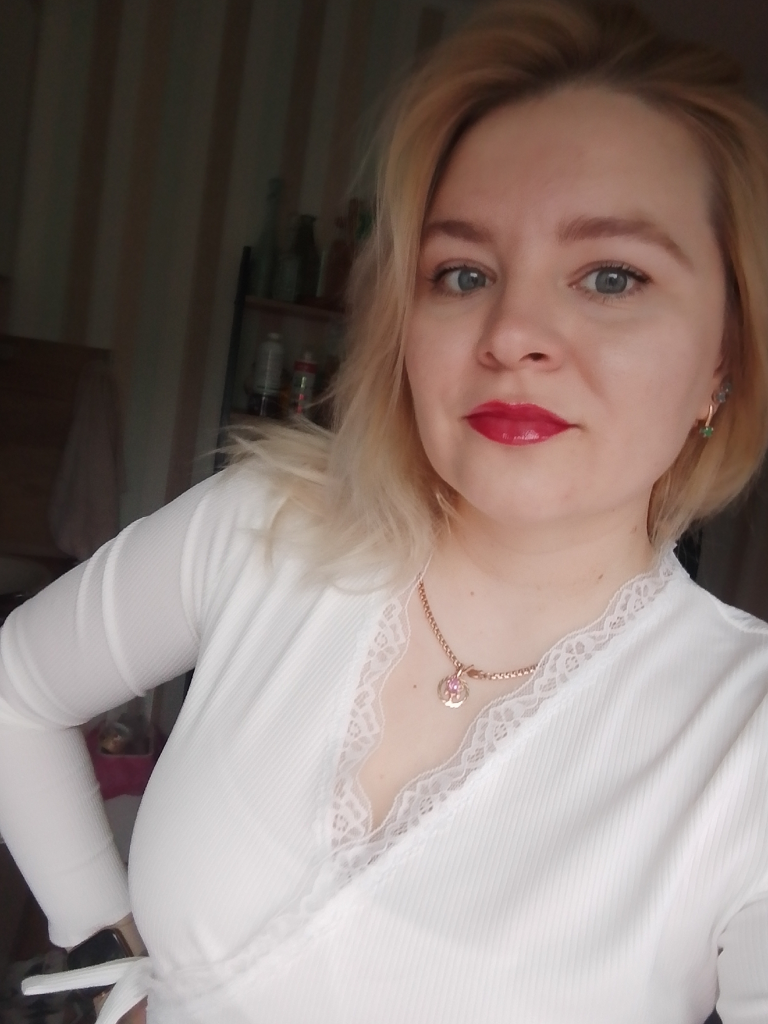 A woman with blonde hair and blue eyes is posing for a selfie. She is wearing a white, long-sleeved, wrap-style top with lace trim at the neckline. A gold necklace with a pendant is visible. She has red lipstick on. In the background, there is a shelf with bottles and containers. The location is likely Izhevsk, Russia, although this is not confirmed by the image.