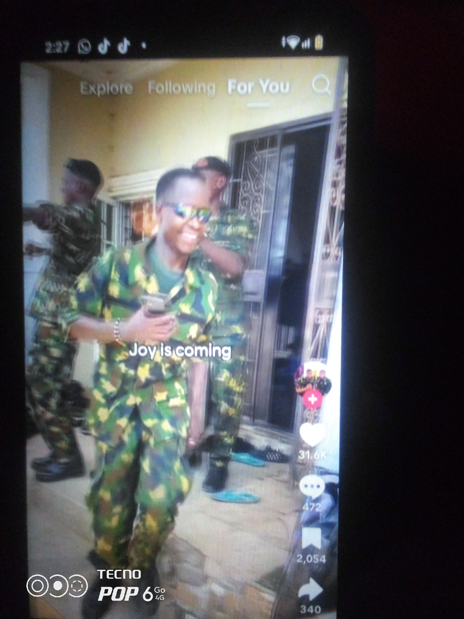 The image shows a screenshot of a mobile phone screen, displaying a TikTok video. The video features several individuals in Nigerian military camouflage uniforms. The main subject in the foreground is a person wearing sunglasses and smiling, holding a clear plastic cup. Text overlaying the video reads "Joy is coming". Other individuals in uniform are visible in the background. On the right side of the screen are typical TikTok icons and engagement metrics, including a heart with "31.6K", a chat bubble with "472", and a bookmark icon with "2,054", and an arrow icon with "340". The top of the screen shows the time "2:27" and other phone status icons, as well as social media navigation buttons "Explore", "Following", and "For You". The scene appears to be outdoors, possibly in front of a building with decorative metalwork on the windows. The branding "TECNO POP 6 4G" is visible at the bottom left of the screen. There is no information in the image that specifies the location as Howayi, Nigeria.