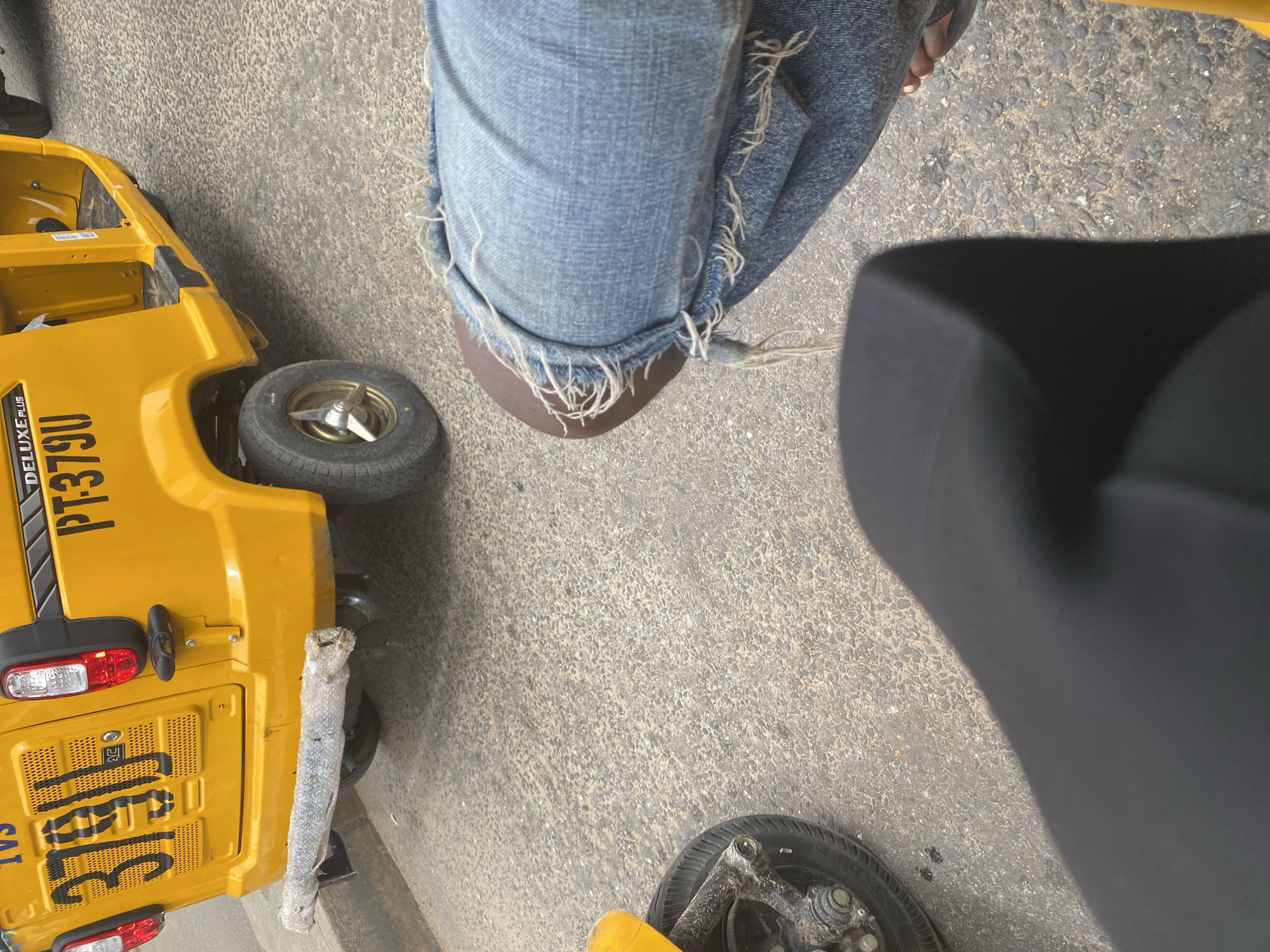 A high-angle shot shows a person's legs in distressed denim jeans, with frayed edges and holes, standing next to a yellow auto-rickshaw. The auto-rickshaw is partially visible, revealing the rear section with a license plate that reads "TVSI 3181G". The words "DELUXE PLUS PT379U" are also visible on the side of the vehicle. A black seat of another vehicle is to the right of the frame. The ground is asphalt, suggesting an outdoor, possibly urban or roadside setting. The lighting suggests it is daytime. The scene appears to be in Jalingo, Nigeria, as indicated by the location context. The image does not provide explicit cues about the weather or any specific event taking place, but the casual attire of the person and the presence of the auto-rickshaw might imply everyday street life or transportation activities.