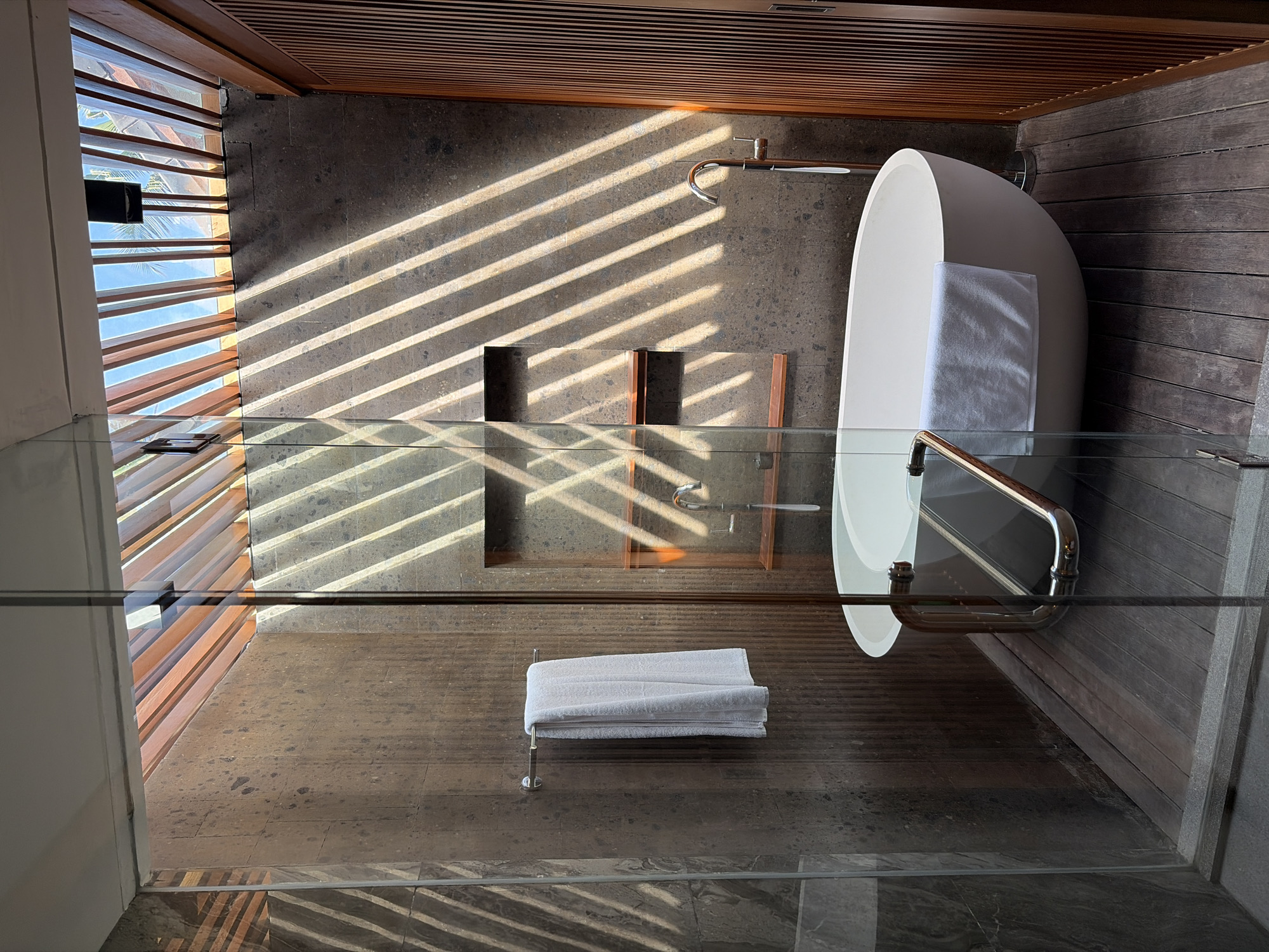 This is a forensic, matter-of-fact description of a bathroom in the Ubud District, Indonesia. The scene depicts an indoor bathroom with a focus on a freestanding bathtub and its surroundings. The bathtub is white and oval-shaped, positioned on a dark wooden slatted floor. A white towel is draped over the side of the bathtub. A glass shower enclosure with a metal handle is partially visible, enclosing the bathtub area. To the left, a silver towel rack is mounted on a textured, dark grey tiled wall, holding a folded white towel. Behind the bathtub, a recessed shelf area is built into the wall, featuring wooden shelves and a chrome faucet. The ceiling appears to be constructed with wooden slats and glass, allowing sunlight to filter through and cast diagonal shadows across the walls and floor. To the right, a wooden slatted door or wall panel is visible. No people are present in the image.