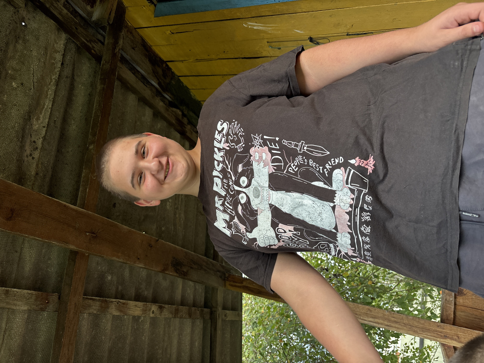 A young person with a shaved head and a bright smile is standing outside under a wooden, unfinished roof. They are wearing a dark t-shirt with a graphic print that features a cartoon character, text, and imagery including a dagger and a skull. The background consists of a yellow wooden wall, greenery from trees, and a glimpse of a blue door or window frame. The overall setting appears to be a rustic outdoor area, possibly a porch or shed.