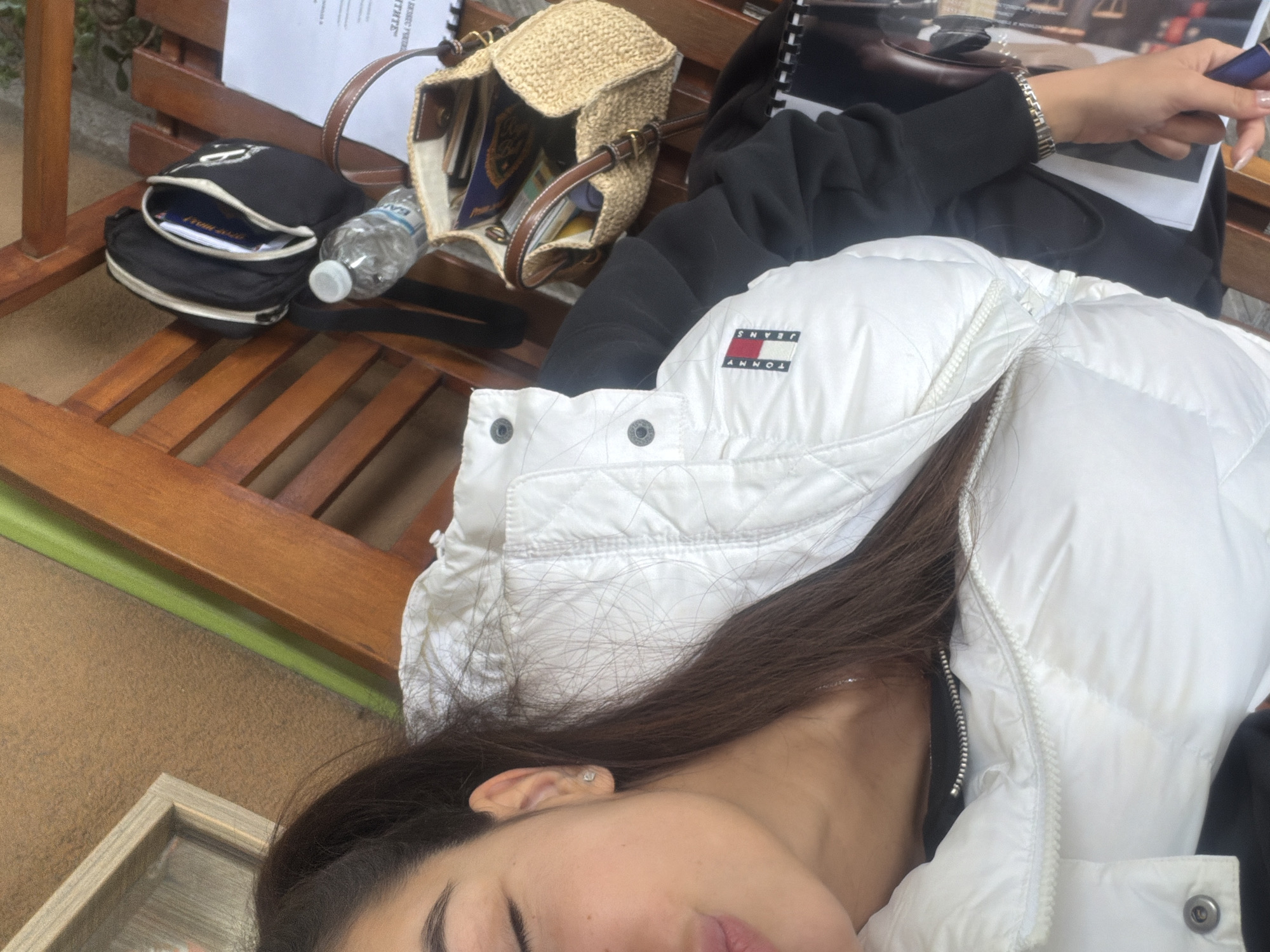 The image shows a partial view of a person lying on a wooden bench. The person is wearing a white puffer jacket with a Tommy Jeans logo. A black zippered bag, a woven handbag, and a plastic water bottle are on the bench beside them. A notebook is also visible next to the person, with a hand holding a pen or marker above it. The location appears to be outdoors. The photograph gives no information to suggest the location is in Plovdiv, Bulgaria.