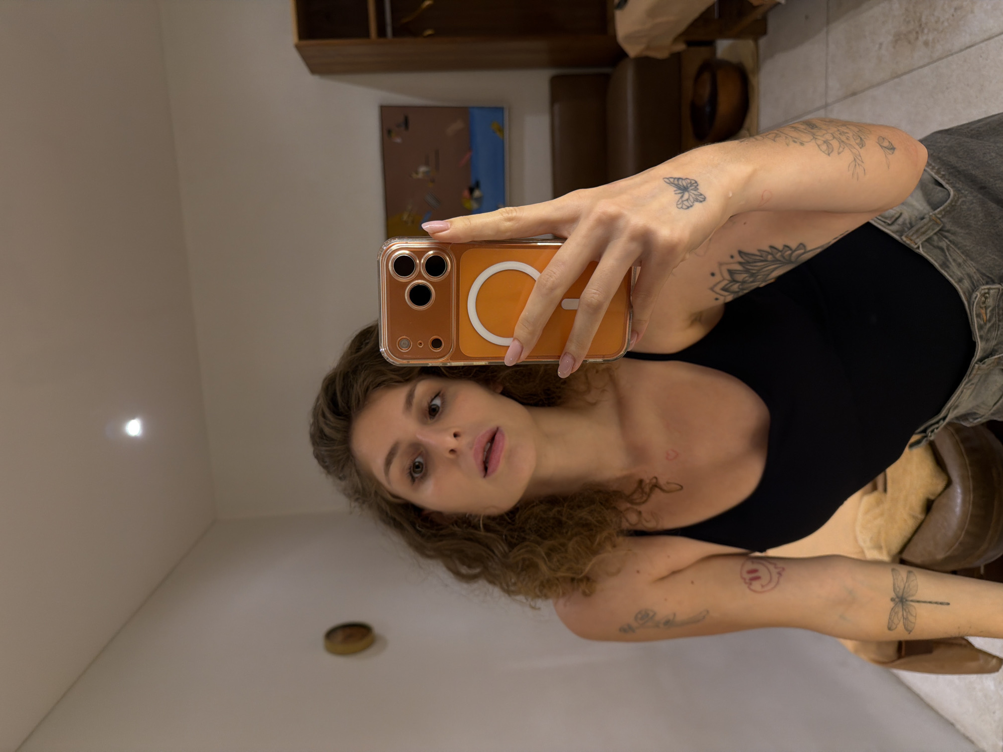 A young woman with curly brown hair and light skin is taking a selfie in what appears to be a hotel room in North Kuta, Indonesia. She is wearing a black tank top and grey denim shorts. She has several tattoos on her arms and chest, including a dragonfly, a butterfly, and a smiley face. She is holding an orange iPhone with a clear case. In the background, there is a bed with tan linens, a wooden cabinet, and a painting on the wall. The lighting in the room is dim.