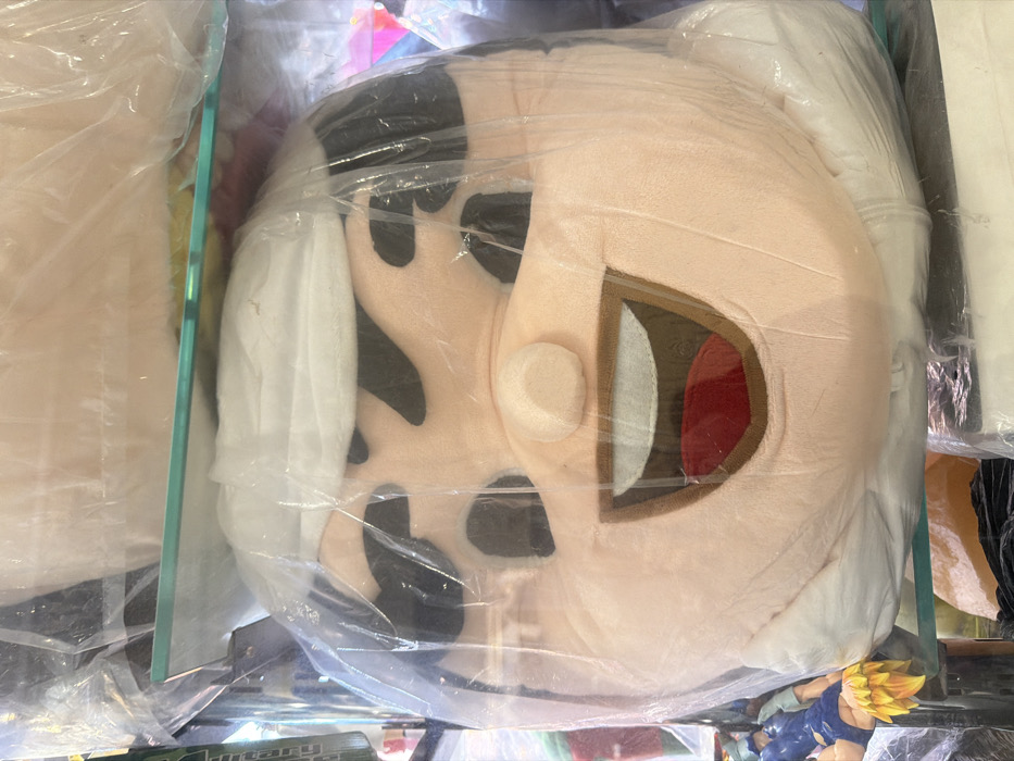 A plush toy of a cartoon character's head rests inside a clear, plastic-wrapped display. The character has pale skin, black hair, and a partially open mouth revealing brown and red colors. A small, round nose protrudes from the face. A glass panel partially separates the plush from other items to the left. A small figurine of a character with yellow, spiky hair is visible at the bottom right corner.