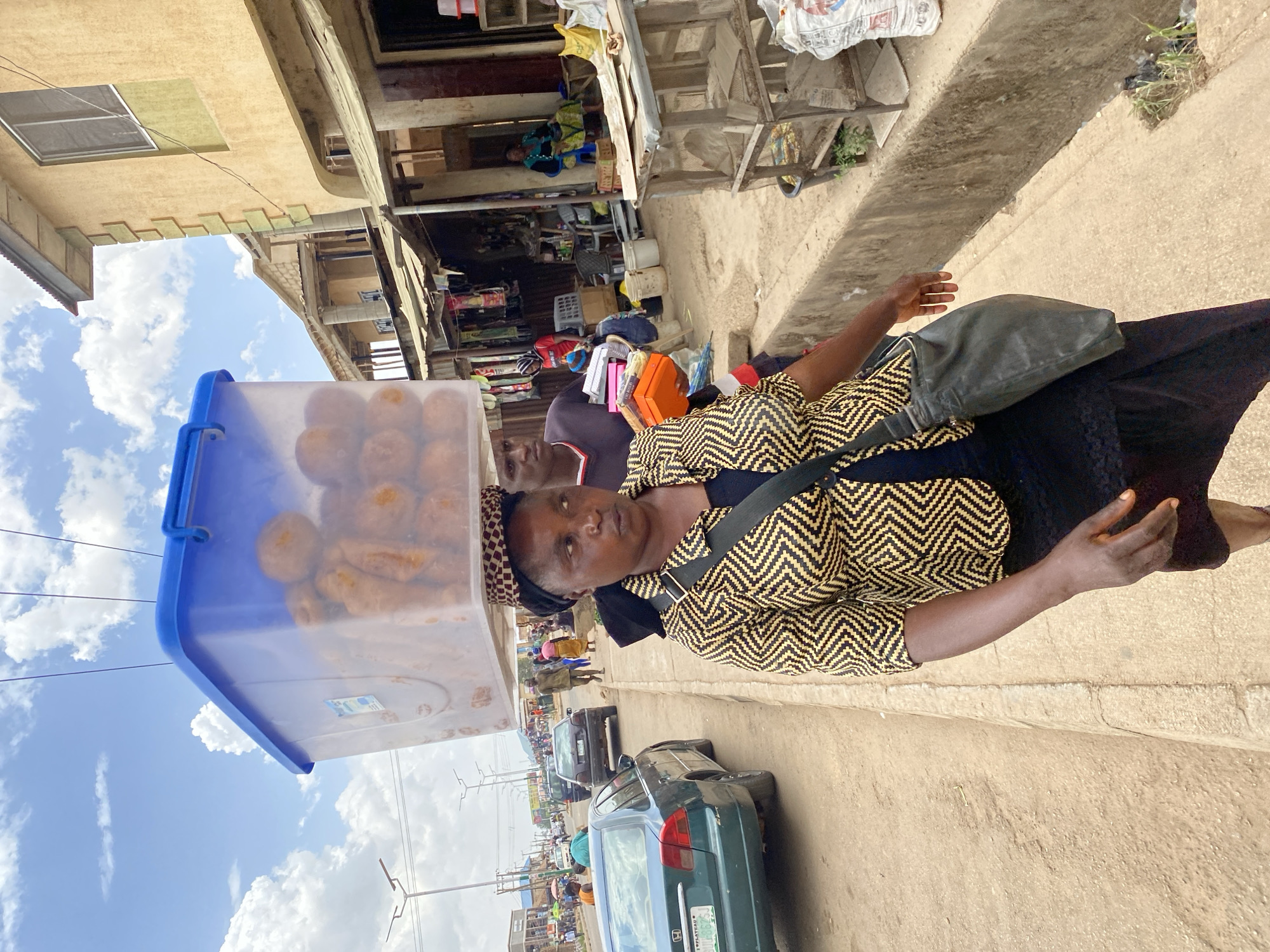A woman stands on the side of a road in Bukuru, Nigeria, carrying a blue plastic container filled with round, golden-brown pastries on her head. She wears a patterned yellow and black shirt, a dark head covering, and a black dress, with a shoulder bag across her body. A man partially visible behind her is also on the road. The street scene shows cars parked along the road and other people further down the street, with buildings visible in the background. The sky above is partly cloudy.