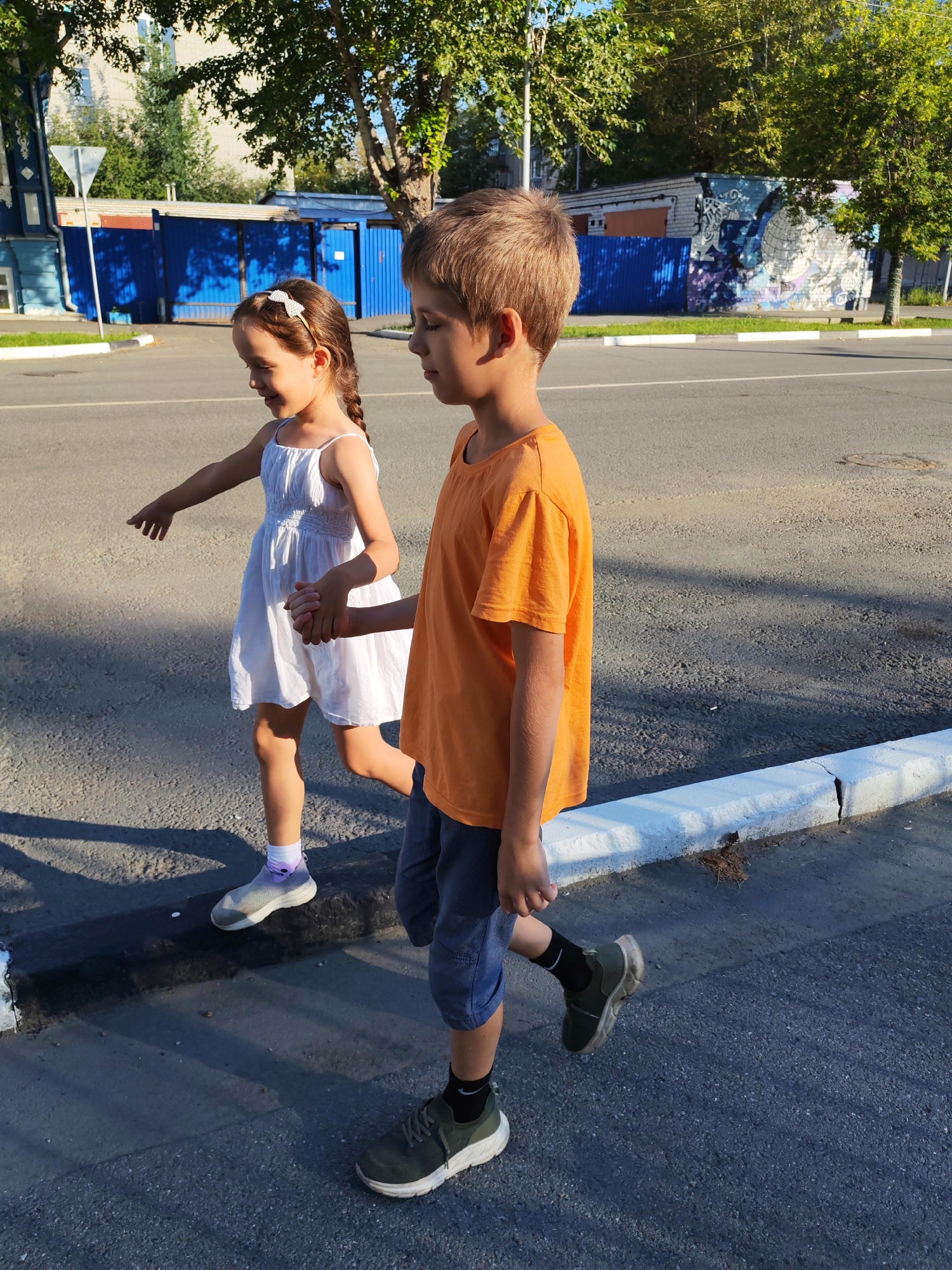 Two children are walking on the sidewalk next to a street in an urban environment. The child on the left is a young girl wearing a white dress and white shoes with purple socks. She has a bow in her hair and is holding hands with the child on the right. She is looking to her left and pointing with her right hand. The child on the right is a boy wearing an orange t-shirt and blue shorts, with dark socks and green shoes. He is looking forward. The background shows a blue fence, a building with graffiti, and trees. The ground is asphalt and the curb is painted white. Sunlight casts shadows on the ground.