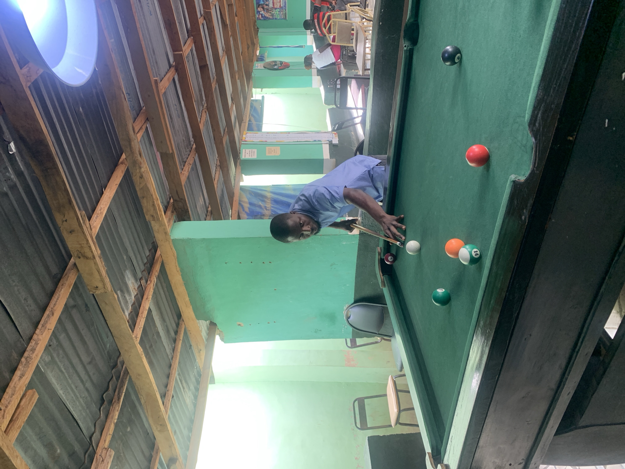 The image shows a man playing pool in what appears to be a pool hall or recreational space. The man is of African descent, wearing a light blue shirt, and is in the process of aiming with a pool cue at the cue ball.
The pool table is green with several pool balls scattered on it, including solid and striped balls, as well as the black 8-ball.
The background shows a sparsely decorated room. The walls are painted light green. Several chairs and people are visible in the background. The ceiling is constructed of corrugated metal supported by wooden beams. A bright light fixture is visible on the ceiling. Based on the mention of New Stadium, Nigeria, it can be assumed that this is the location, though no explicit landmarks confirm this.