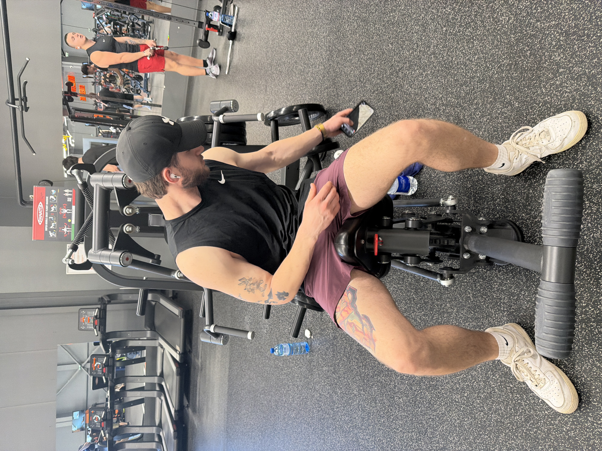 The image depicts a man sitting on a gym machine in what appears to be a fitness center. He is wearing a black sleeveless shirt, maroon shorts, a black baseball cap, and white sneakers with socks. He has a beard and tattoos on his arms and right thigh. He is holding a smartphone in his right hand. A water bottle is visible on the floor near the machine. The gym has mirrors, weight machines, treadmills, and other fitness equipment.
It is not possible to confirm if the location is in Plovdiv, Bulgaria based on the image alone. Additional information would be needed to determine the specific location.