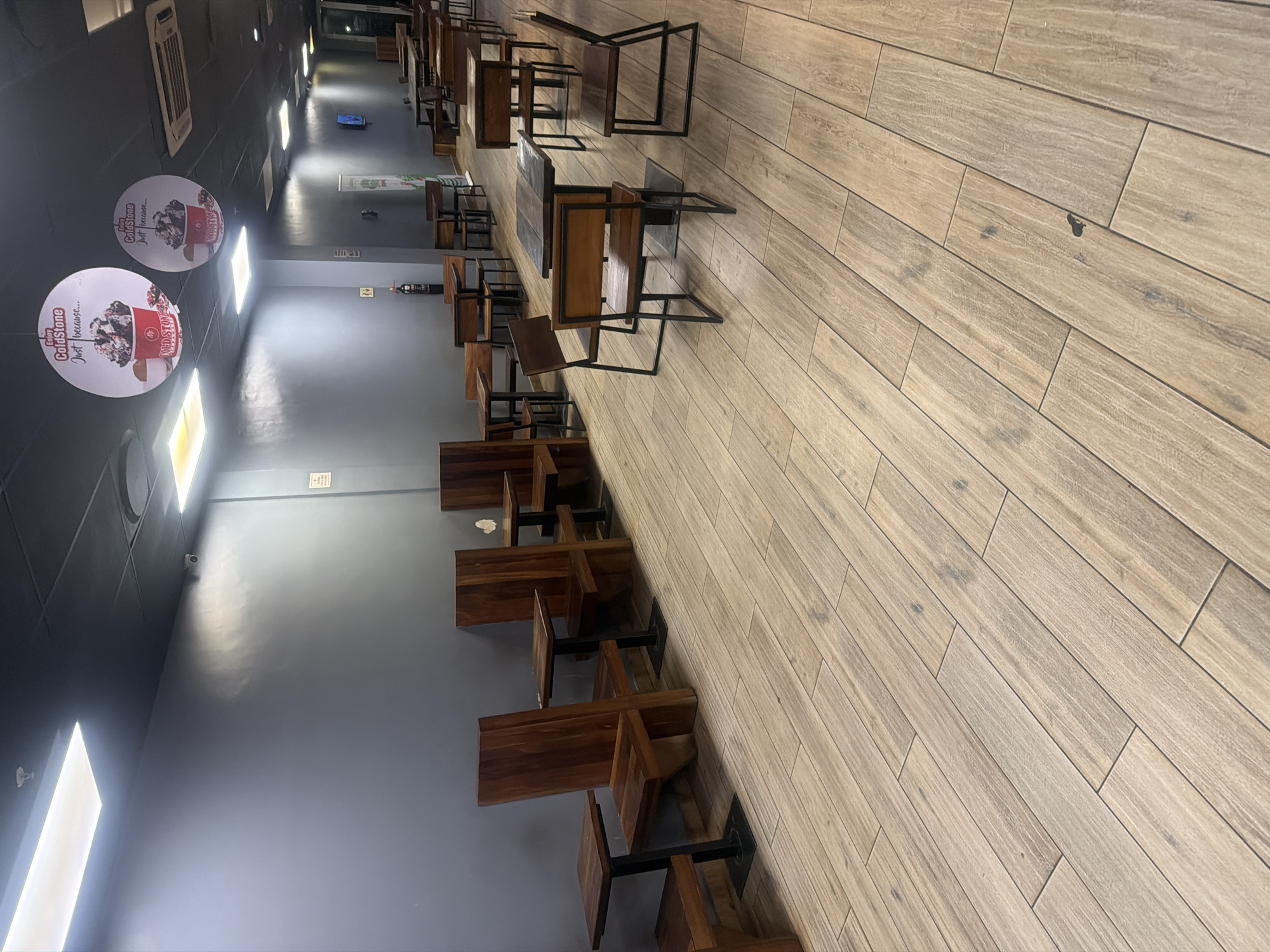 This media file captures the interior of a commercial dining establishment, likely a cafe or restaurant, located in Abuja, Nigeria. The scene depicts a long, rectangular room with a dark grey-blue painted wall on the left and a light-colored, wood-look tiled floor extending to the right. The ceiling is dark, featuring numerous recessed rectangular light fixtures and an air conditioning unit.
The space is furnished with many wooden tables and chairs, each having black metal frames. These are neatly arranged in rows along the left wall, with chairs either pushed under the tables or partially stacked, indicating the venue is currently empty or closed.
Visible branding includes two large, circular promotional signs for "Cold Stone Creamery" hanging from the ceiling. Both signs display the text "Enjoy ColdStone Just because...". Other notable objects include a small security camera mounted high on the wall, a fire extinguisher, and a white sign on the wall with "NO SMOKING" printed on it. Further down the aisle, a blue object, possibly a cleaning cart or bucket, is visible on the floor.
No people are present in the image, and no discernible activity is taking place. The artificial lighting inside means the specific time of day is indeterminate. The overall impression is one of stillness in an unoccupied commercial space.