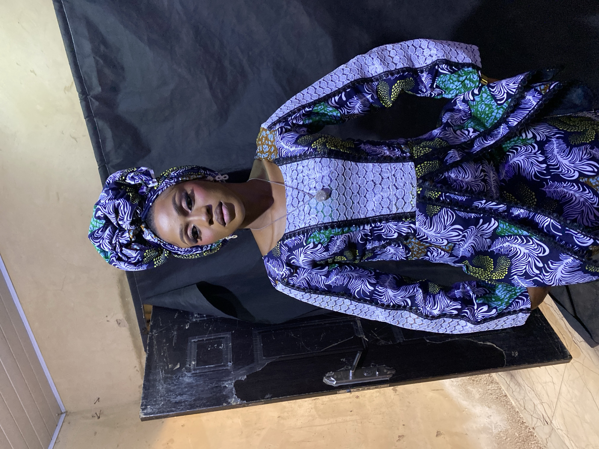 A formal portrait captures a young woman looking directly at the viewer with a calm, neutral expression. She is adorned in a vibrant, patterned outfit, likely an Ankara-style print, featuring a dark blue base with intricate swirls of white, light blue, green, and yellow. A matching, intricately tied headwrap (gele) frames her face. The dress incorporates panels of white lace on the bodice and sleeves, adding a touch of elegance. She wears a delicate silver chain necklace with a small, light-colored pendant. Her makeup is well-applied, with noticeable eyeshadow and lip color enhancing her features.
The setting appears to be an indoor space, possibly a makeshift studio, suggested by the dark, wrinkled fabric draped behind her as a backdrop. Below her, a dark, worn surface, resembling a table or cabinet, provides a foreground element. The periphery of the image reveals a lighter, beige-colored wall or floor, showing some signs of wear. The lighting is artificial, accentuating the details of her attire and facial features. The photograph was taken in Kuru Dungus, Nigeria. There is no visible text in the image.
