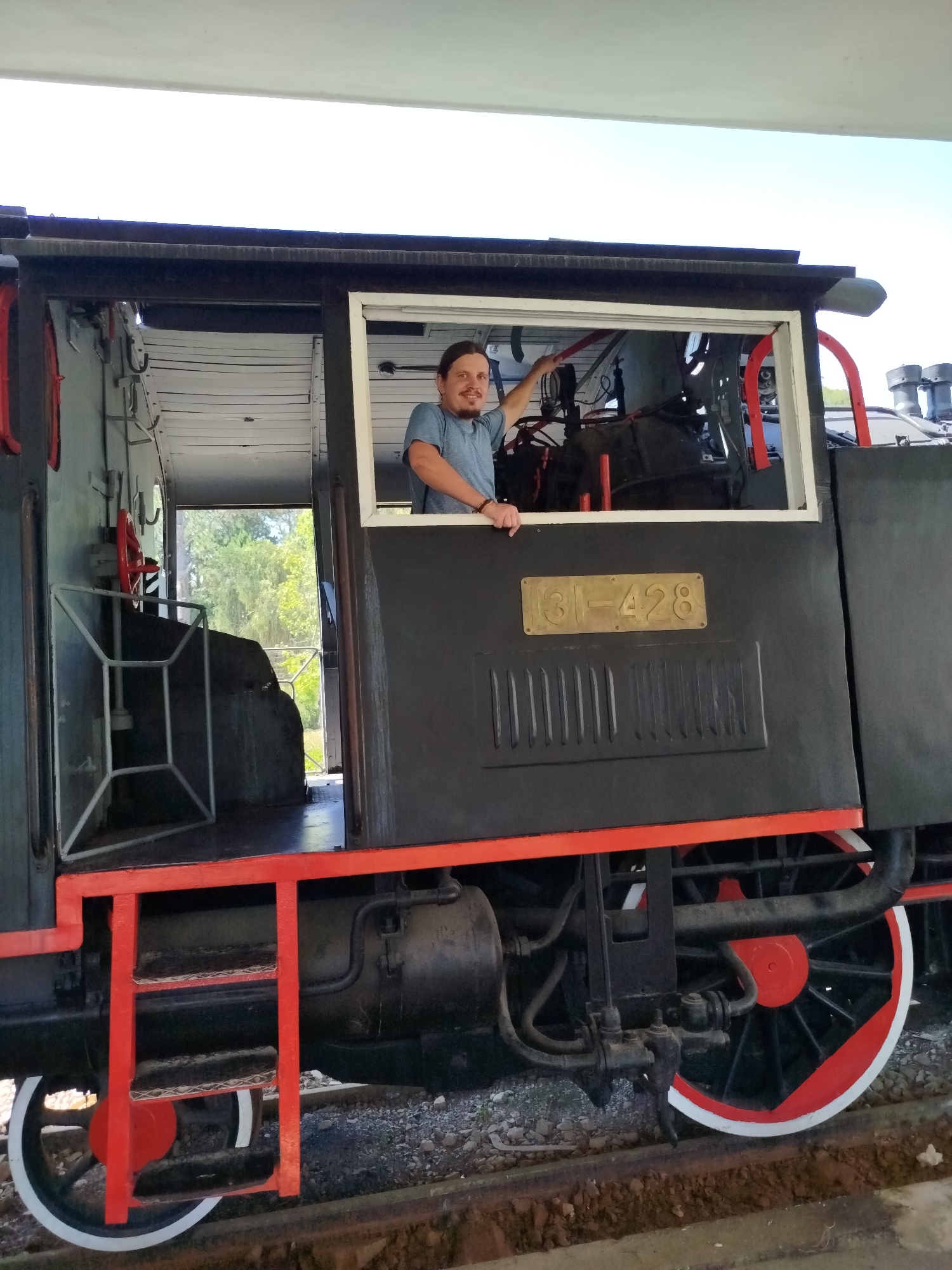An adult male is positioned within the cab of a black vintage steam locomotive, identified by a brass plaque displaying "31-428," located at the Đà Lạt Railway Station in Thành phố Đà Lạt, Vietnam. The man, with long dark hair, a beard, and a mustache, wears a light blue short-sleeved t-shirt and a braided wristband on his left arm. He is leaning out of a rectangular window opening on the right side of the cab, with his left forearm resting on the window sill and his right hand near an internal red lever. He is looking towards the viewer with a smile.
The locomotive features red accents on its steps and a portion of its wheels, which are also partly white. The cab interior reveals a white slatted ceiling, various controls, a red handwheel, and a light-colored, geometric metal frame to the left. The locomotive rests on train tracks, with gravel and soil visible beneath. A large, light-colored overhead canopy provides shelter above the scene. Through an opening at the rear of the cab, lush green foliage is discernible, indicating an outdoor area beyond the locomotive's immediate display location.
