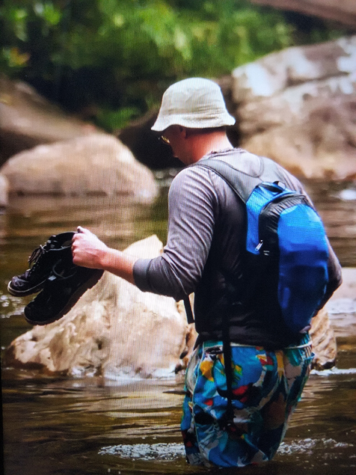 A person is walking through shallow water with a backpack and holding a pair of boots. The person is wearing a grey long-sleeved shirt, a light-colored bucket hat, and brightly colored shorts. The backpack is blue and black. The water is murky and reflects the surrounding environment. Large, light-colored rocks are visible in the water. In the background, there is lush green foliage. The scene appears to be in a natural, outdoor setting, likely a river or stream. The location is not specified as being in the city of Payagaboh, Indonesia, or any other city.