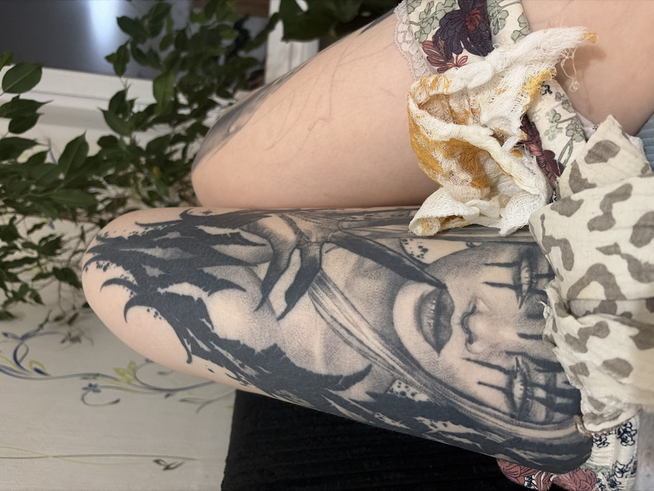 An upper arm bears a detailed tattoo of a woman's face with markings beneath her eyes, rendered in shades of gray and black. The arm rests atop another, also displaying a faint tattoo. A floral-patterned garment with lace trim adorns the upper arm, intertwined with textured fabrics in cream, yellow, and muted animal print. Foliage is visible in the background.