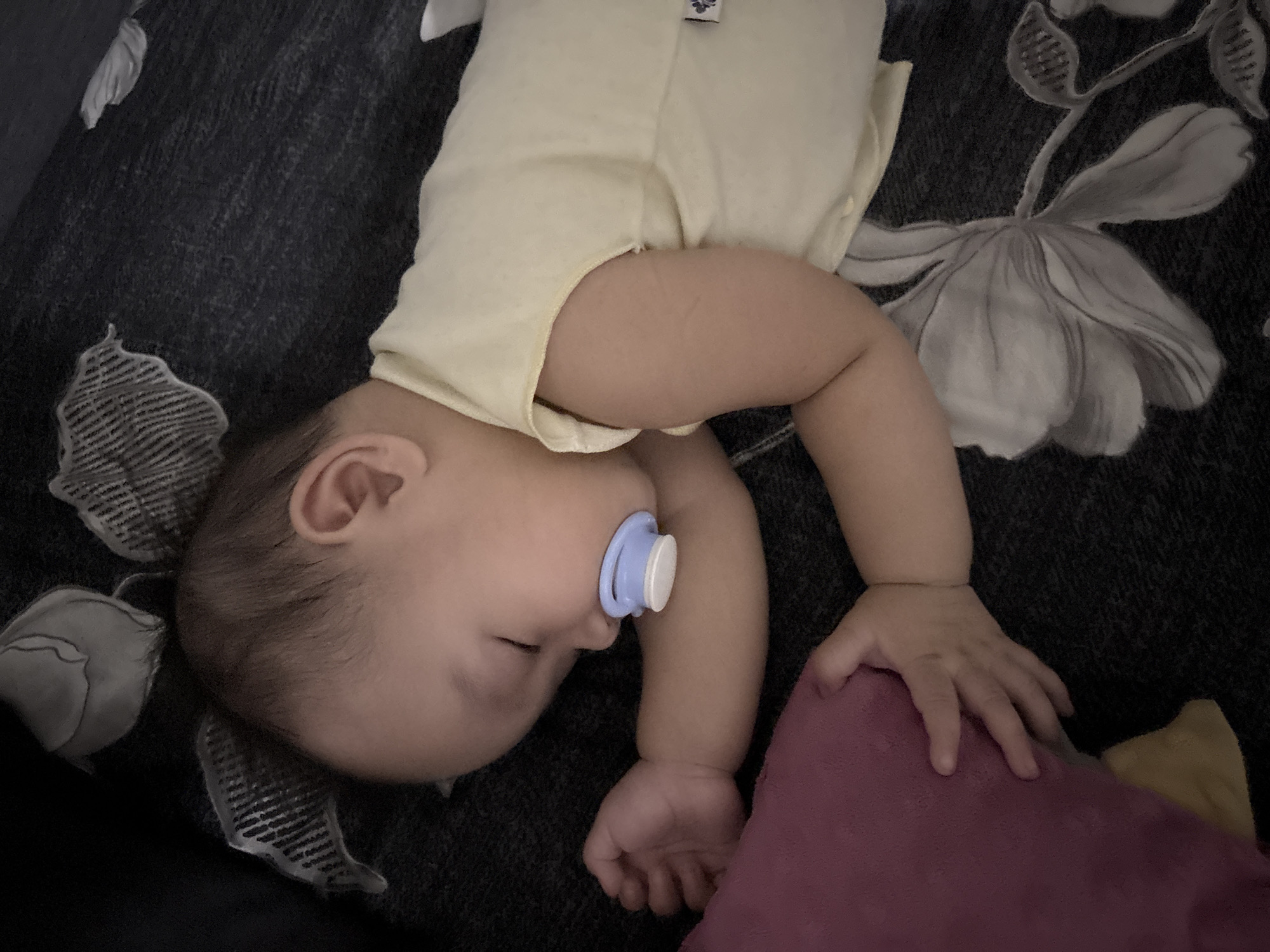 A baby is sleeping soundly on a dark, floral-patterned surface, possibly a bed or couch. The baby is wearing a light yellow sleeveless onesie and has a light blue pacifier in its mouth. The baby's right arm is resting near a plush pink object, likely a pillow. The overall lighting suggests an indoor setting. This image does not provide any specific location in West Jakarta, Indonesia.