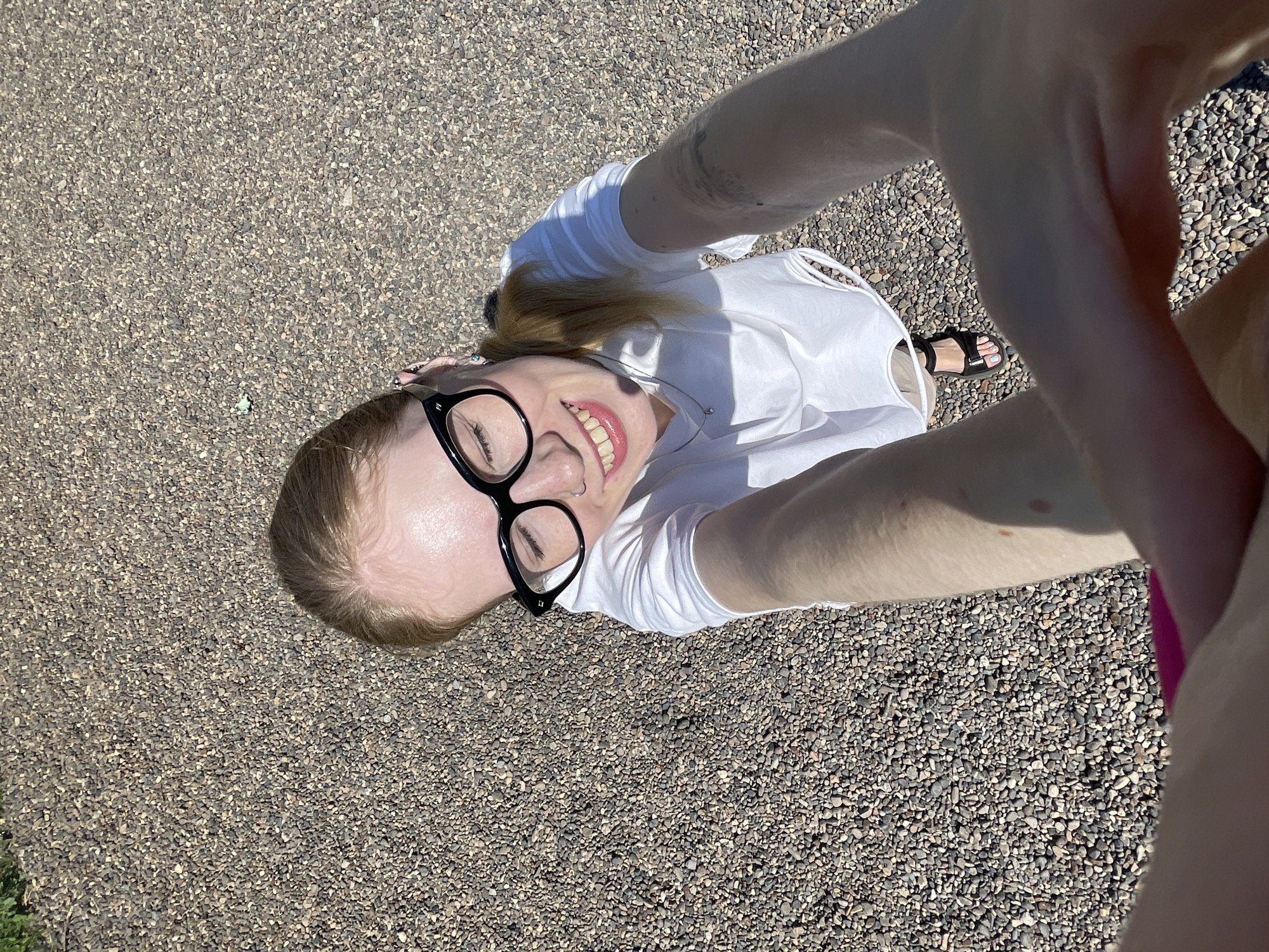 A woman with blonde hair and black glasses is taking a selfie outdoors on a pebbled surface. She is wearing a white top and black sandals. A pink garment is visible on the right side of the frame. The location appears to be a paved area. The location is not verifiable as Izhevsk, Russia based on the visual information provided.