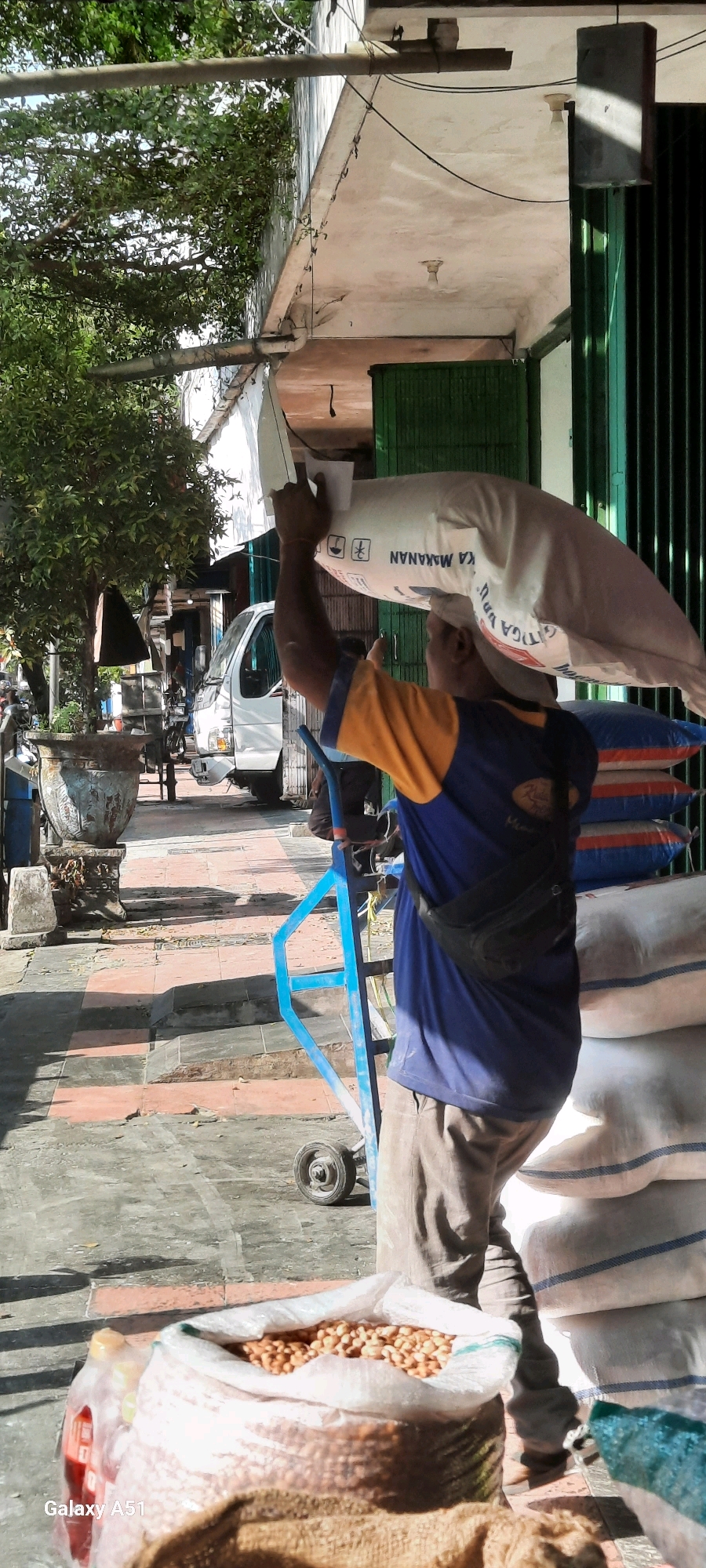 An outdoor scene in Beru, Indonesia, captures a man carrying a large sack of food products on his head. The man, wearing a blue and yellow shirt and grey pants, is walking on a paved sidewalk next to a building. To his right, a stack of white sacks with blue stripes are visible. The background shows a white truck, a large decorative pot, and greenery from trees. The sunlight casts shadows on the ground, indicating it's daytime.