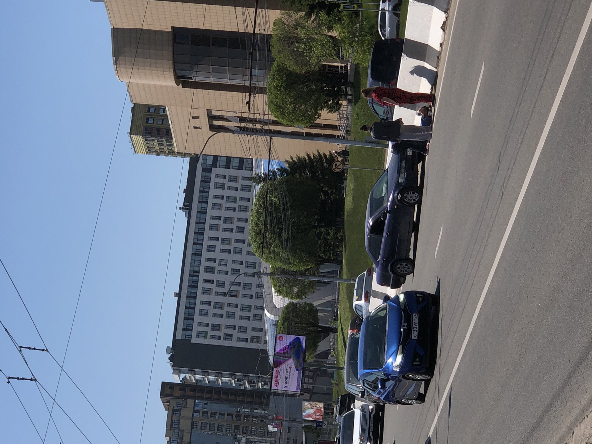 This image captures a bright, sunny urban street scene in Krasnoyarsk, Russia. The view looks down a multi-lane asphalt road with clear white markings, extending from the foreground into the distance. To the left of the road, there's a sidewalk, a curb with black and white markings indicating a pedestrian crossing, and a grassy verge lined with trees and utility poles. Beyond this, several multi-story buildings of varying architectural styles form the backdrop, under a clear blue sky crisscrossed by power lines.
The scene depicts everyday city life during daylight hours. Several cars are parked along the curb on the left side of the road. In the foreground, a prominent blue Honda (likely a Fit or Jazz model) displays the Russian license plate "K133YB124," with the "124" region code identifying Krasnoyarsk Krai. Further down the street, other dark-colored sedans and a silver car are visible.
A few individuals are present near the parked cars on the sidewalk. One person, wearing a red patterned outfit, stands near a dark car, with another person in a dark top and grey pants standing nearby. Their activity appears to be casual, possibly conversing or waiting.
Notable details include a clear blue sky, indicating good weather. A street sign with a red circle and a diagonal line (often signifying "No Stopping" or "No Parking") is visible in the middle ground. A large digital billboard advertises "4K ВИДЕО" (4K VIDEO), "XIAOMI 11T," and "М.ВИДЕО" (M.Video, a Russian electronics retailer), further confirming the Russian location. Another smaller billboard is also visible below it. The overall impression is a typical bustling city street on a clear day.