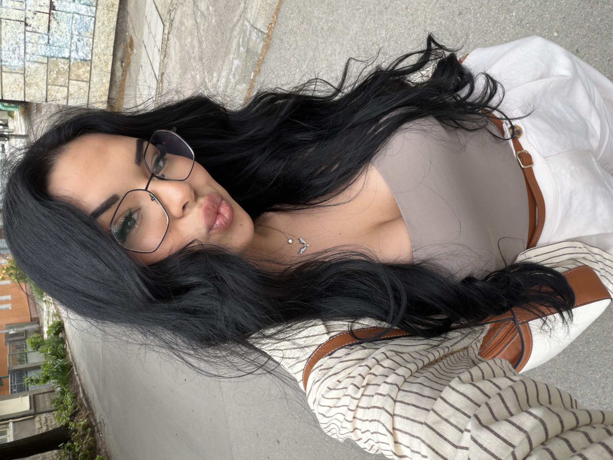 A woman is seen in a selfie taken outdoors, with an urban environment visible in the background. She has dark hair, wears glasses, and is dressed in a neutral-toned top, white clothing, and a striped jacket. A necklace is visible around her neck. The background shows an urban setting with buildings, a street, and some greenery, implying the city of Unknown, Unknown.