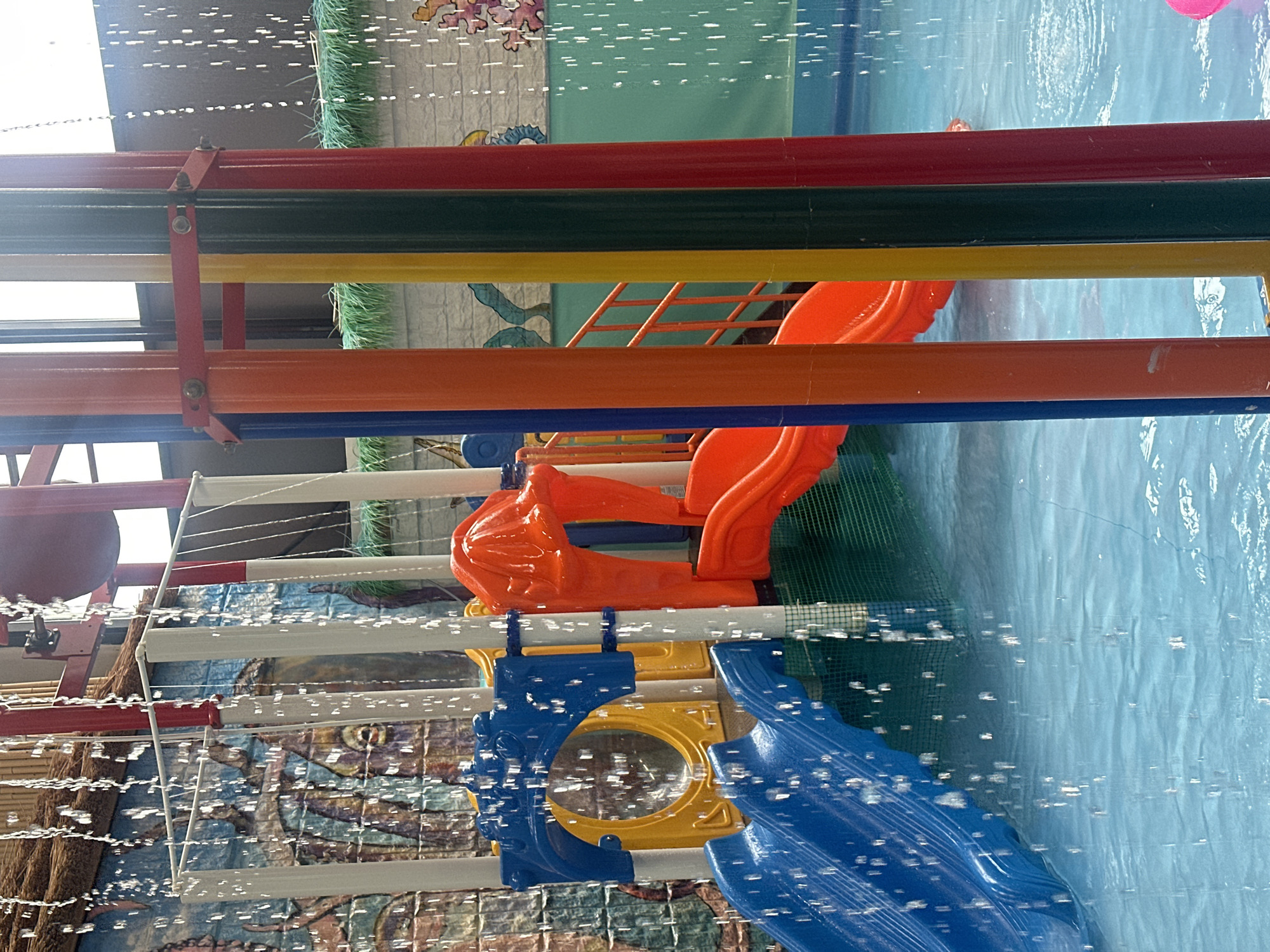 The image depicts a colorful water park playground structure with slides and water features. The playground is made of bright primary colors like blue, orange, yellow, and red. Water is cascading down in streams, creating a lively and dynamic scene. In the background, there appears to be a mural or decorative wall with ocean-themed imagery. The entire structure is situated in a pool of clear blue water. The location is the "Kva-Kva Park" water park, located at 18-ya Myasnitskaya Street, building 1, in Moscow, Russia.