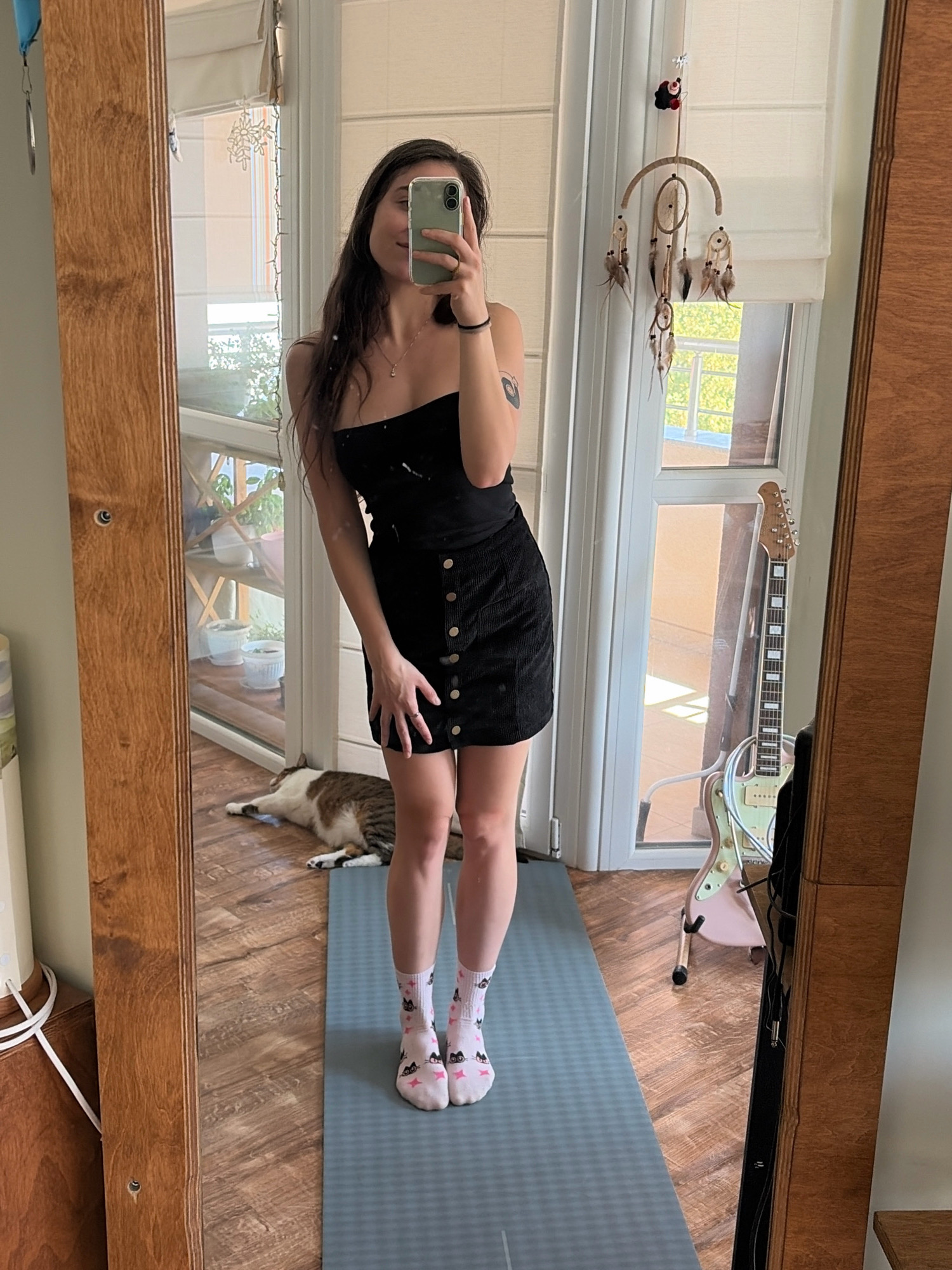 A young woman is taking a mirror selfie in a room. She is standing on a blue yoga mat, wearing a black strapless dress and white socks with cat patterns. She is holding a phone in her right hand and wearing a ring on her left ring finger. In the reflection of the mirror, a tabby cat is lying on the floor, and a light green electric guitar with a pink stand is visible. The room has wooden flooring and a wooden mirror frame. A dreamcatcher hangs on the wall near a window with white blinds.