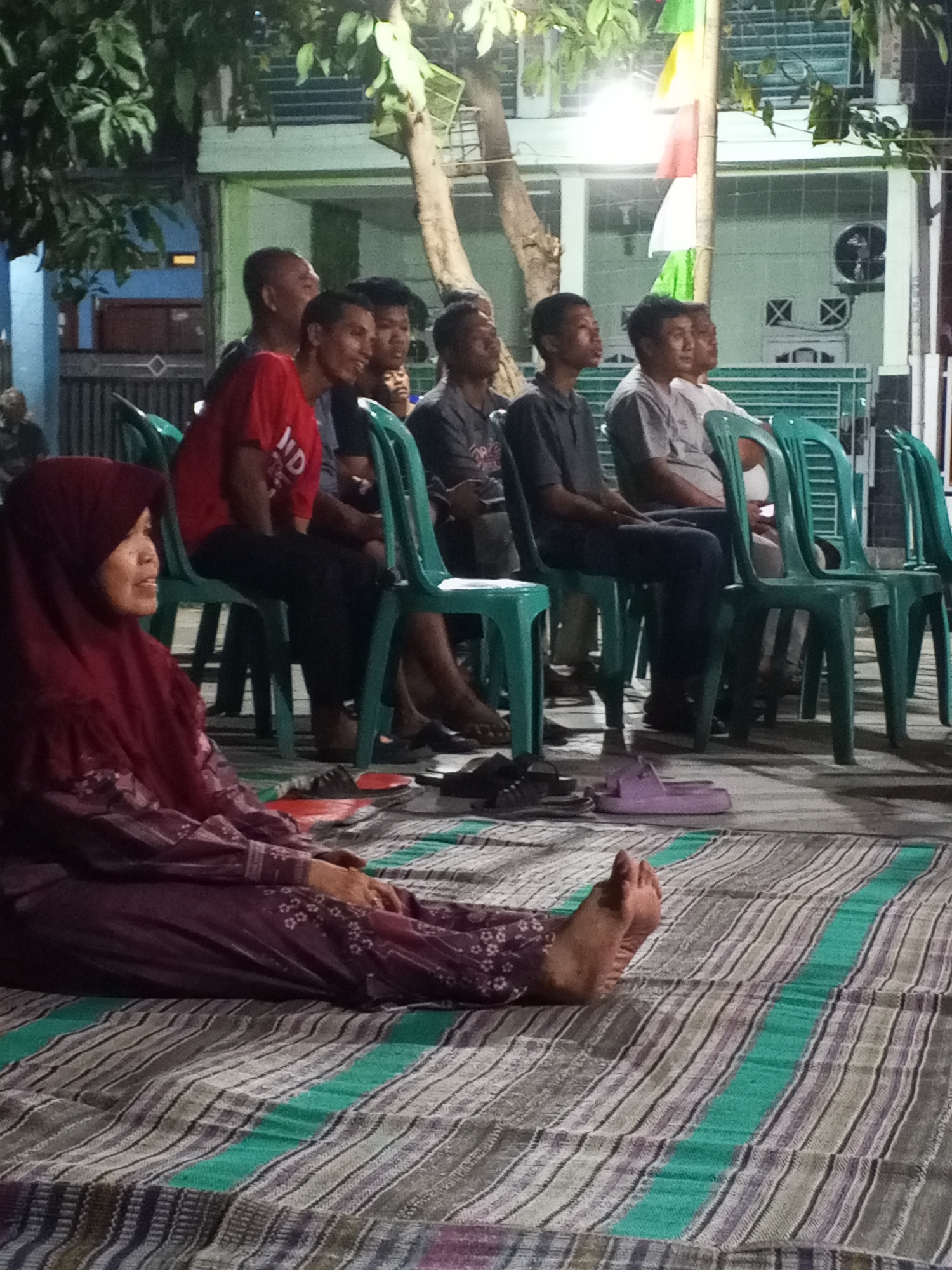 The image shows a group of people sitting in chairs and on the ground, possibly attending an outdoor event. In the foreground, a woman wearing a maroon hijab and a patterned dress is seated on a striped mat. Her feet are visible, with one foot slightly raised. To her right, several men are seated in green plastic chairs. One man in a red shirt is looking to the left, while another in a dark gray shirt is looking forward. In the background, there are buildings with windows and a tree with green leaves. The overall setting appears to be an informal gathering in a residential or community area, possibly in Ciruas, Indonesia, given the context.