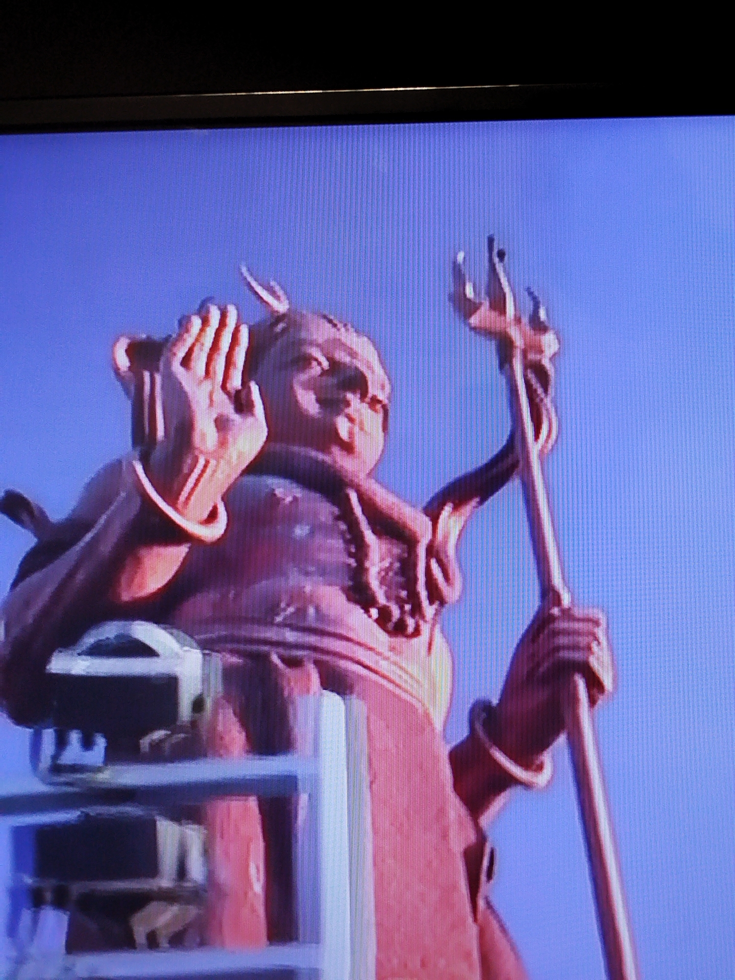 A statue, possibly of a deity or mythological figure, is shown against a clear blue sky. The statue is made of a reddish-brown material and appears to be holding a trident in its right hand. The figure has a large head with prominent features and is wearing a necklace. Part of what appears to be scaffolding or a construction structure is visible in the lower left portion of the image, supporting the statue. The statue is located in Naberezhnye Chelny, Russia, specifically in the area of the Khram Svyatoy Troitsy (Holy Trinity Church) complex, where the statue of the Prophet Elijah is situated.