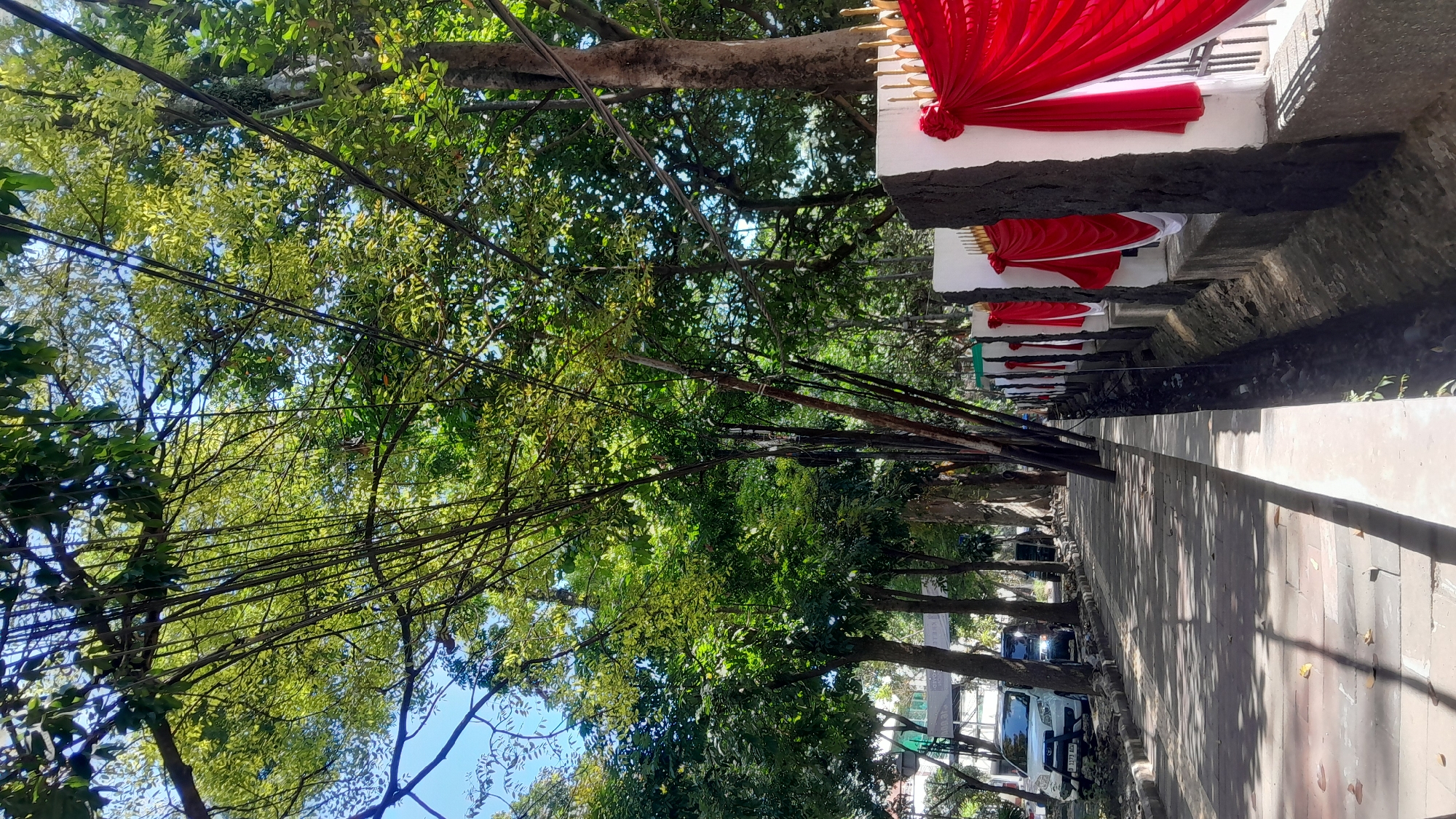 The image captures a street scene with a paved sidewalk on the left and a decorative wall on the right. Tall trees with lush green foliage line the street, casting dappled shadows on the sidewalk. The wall on the right is adorned with red and white drapes, suggesting a festive occasion or a public space decorated for an event. Several poles, possibly temporary structures or natural elements from the trees, are visible. The sky is a clear blue. In the background, cars are parked along the street. Based on the visible license plate format (e.g., "B 1173 AGH"), the location is likely in Indonesia, and more specifically, given the context of other images, this scene is in Bandung City, Indonesia.