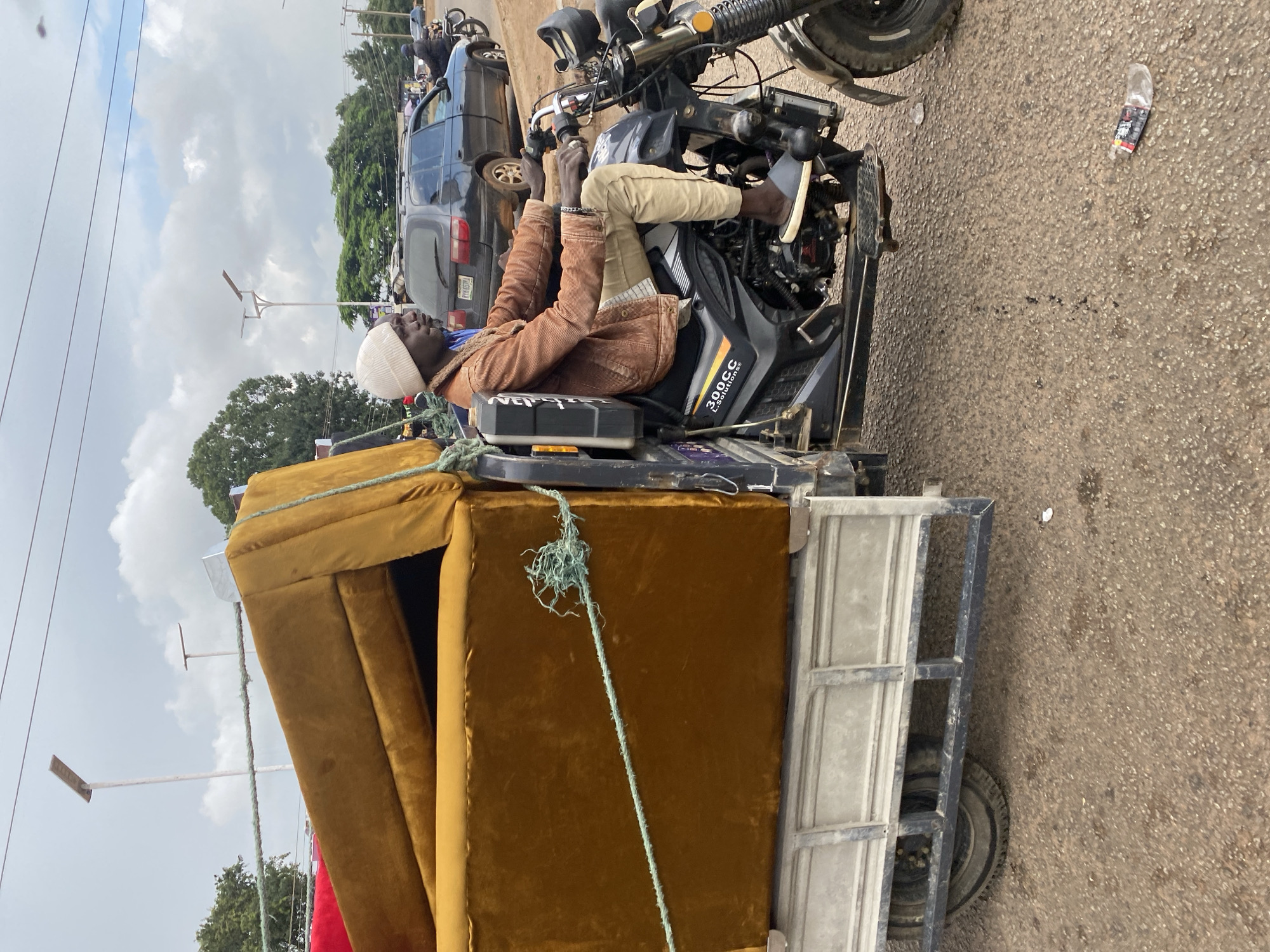 An adult male wearing a light-colored beanie, a brown corduroy jacket, and khaki pants is driving a three-wheeled motorcycle with a trailer. The trailer is loaded with a large, golden-brown sofa and a portion of a red sofa. The motorcycle has the label "300CC L.Solutions" on its side. The man is looking ahead and his hands are on the handlebars. He is wearing gray slippers. In the background, a dark-colored car is parked, and further back, trees and buildings are visible. The ground is paved with asphalt. The scene appears to be on a road in Bukuru, Nigeria, under a cloudy sky with power lines overhead.