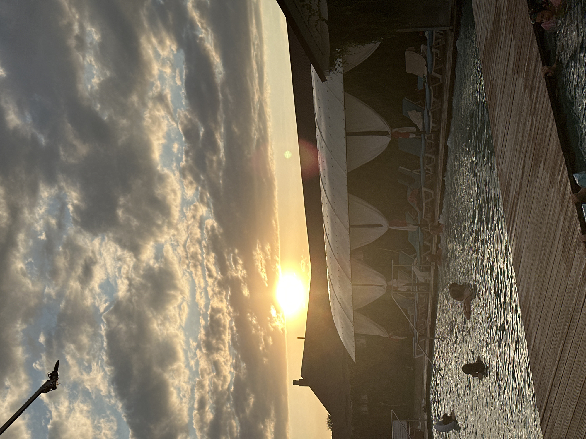 An outdoor swimming pool area is depicted at sunset, with the sun prominently visible in the sky behind a building's rooftop. The sky is filled with dramatic clouds, tinged with the warm hues of the setting sun. In the foreground, a wooden deck leads to the pool, where several individuals are swimming and relaxing. The pool area is furnished with lounge chairs and covered cabanas. A lamppost is visible in the upper left corner of the image, extending from off-frame. The scene is set in Yaroslavskaya, Russia.