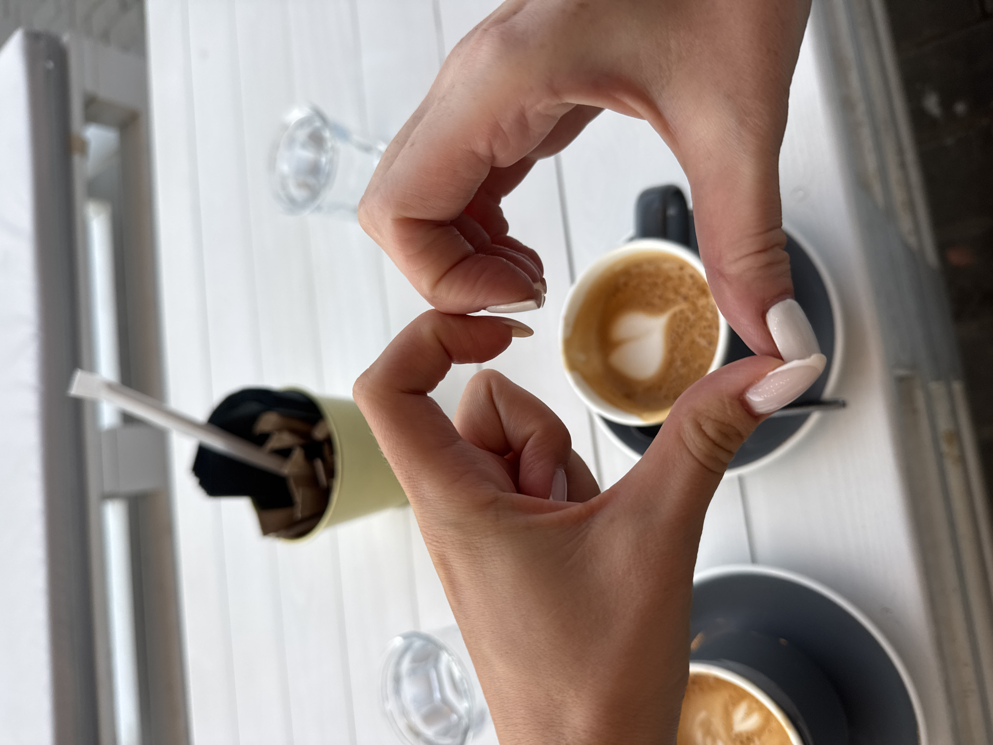 The image presents a top-down, slightly angled view of a person's hands framing a coffee cup on a light-colored slatted table. Two hands, appearing to be female due to their shape and manicured nails with a light polish, are positioned to form a heart shape with their fingers, centered around one of the coffee cups.
On the table, there are two cups of coffee, each with latte art featuring a heart design in the foam. These cups are white with dark grey or black saucers. Near the coffee cups, two empty clear glasses are visible. A light green cylindrical container, filled with what appear to be stirrers or sugar packets, is also on the table.
The setting appears to be a bright, well-lit environment, likely a cafe table, possibly outdoors or by a large window, given the natural light and the visible blurred elements suggesting a building or structure in the background. The table surface is made of light-colored, possibly white, slats. The activity captured is an artistic pose, typically for a photograph, highlighting the coffee and expressing affection or appreciation through the heart-shaped hand gesture. There is no visible text in the image. The time of day is clearly daytime due to the bright, natural illumination.