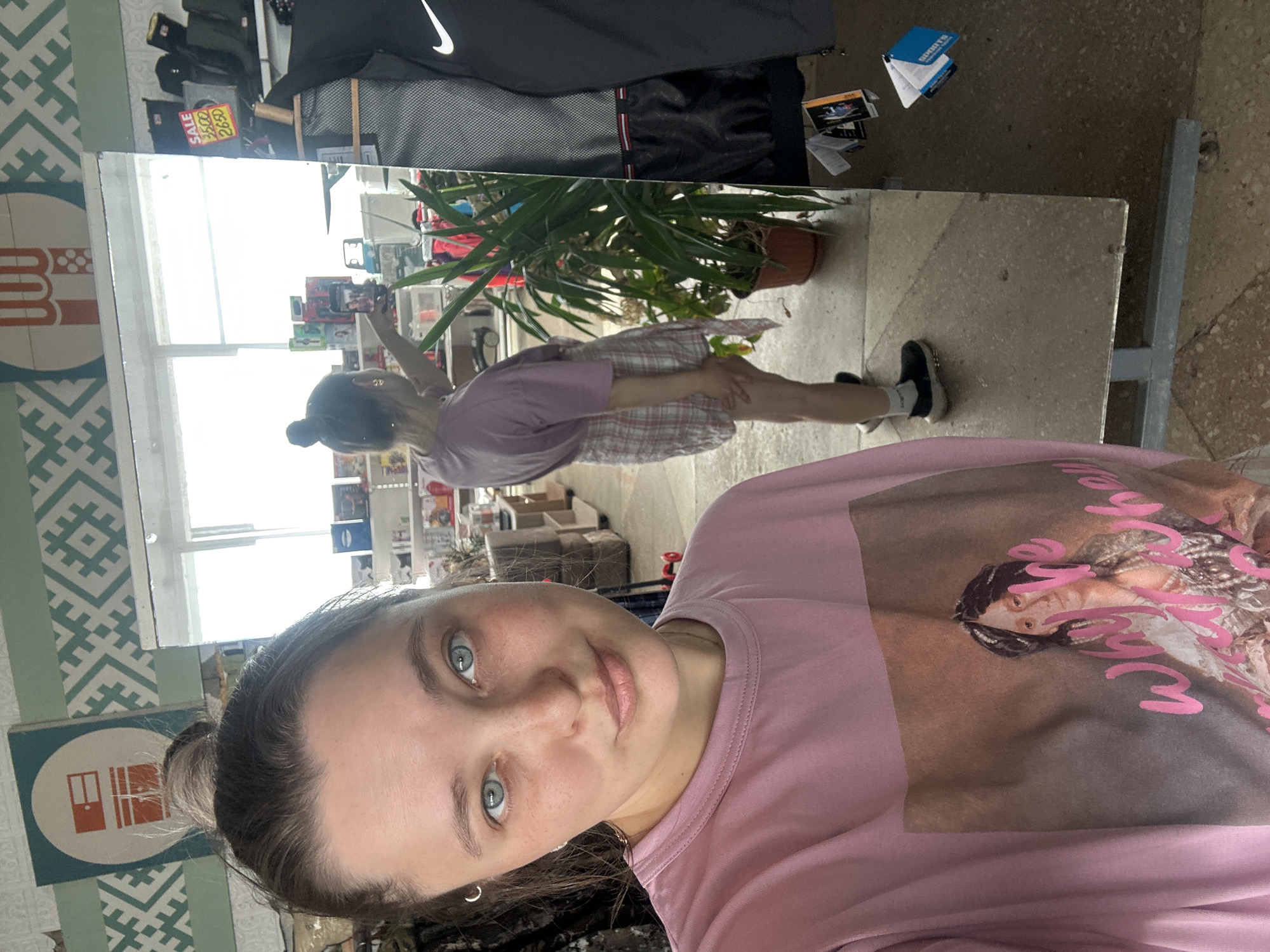 This media file captures a woman taking a mirror selfie in what appears to be a retail environment. The woman, positioned in the foreground, has light skin and blue eyes and is wearing a pink t-shirt with a graphic print. She has her hair pulled back into a bun. In the reflection of the mirror, another woman is visible, also taking a photo with her phone. This second woman is wearing a pink t-shirt and a plaid skirt. She is standing near a potted plant and shelves stocked with various items. Behind her, a large window offers a view of the outside, suggesting it is daytime. The background of the mirror's reflection shows more shelving and merchandise, indicating a store setting. The overall mood appears casual and everyday. Text is visible on a sign in the reflection reading "SALE 3500 2650" and on a blue and white brochure on a surface.