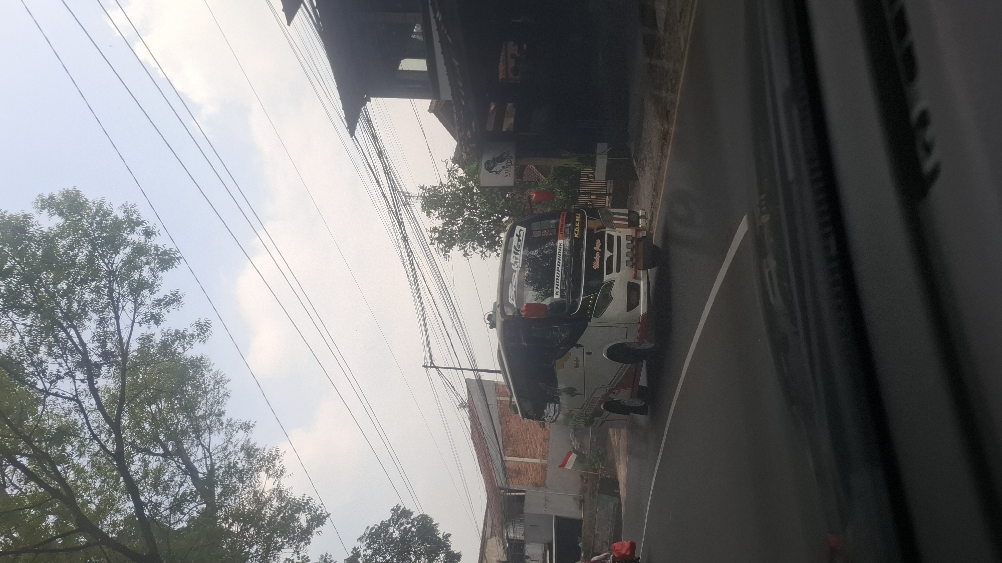 A white and black bus with red accents is driving on a road in Cilaku, Indonesia. The bus has "KADUPANDAK" written on its front. There are buildings and trees on the side of the road, and power lines are visible overhead. The sky is cloudy.