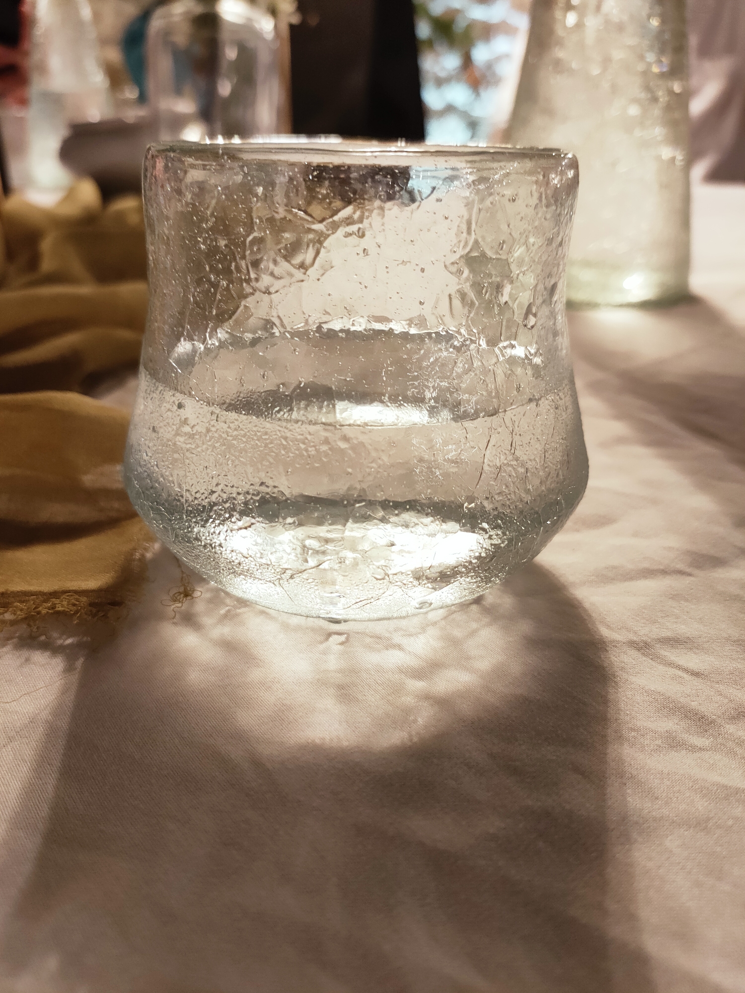 A close-up, eye-level shot captures a textured glass tumbler filled with clear liquid, resting on a light-colored, creased fabric surface. The glass exhibits a rough, uneven surface with visible imperfections and air bubbles, and the liquid inside shows distinct horizontal layers. A folded piece of golden-brown fabric is positioned to the left of the glass, partially obscuring the background, which is blurred and out of focus, showing hints of other glassware and dimly lit objects. A distinct shadow of the tumbler is cast onto the fabric beneath it. The scene appears to be set indoors, possibly at a dining or event table, with soft lighting creating a warm ambiance. The photograph is taken in Cebalat, Tunisia.