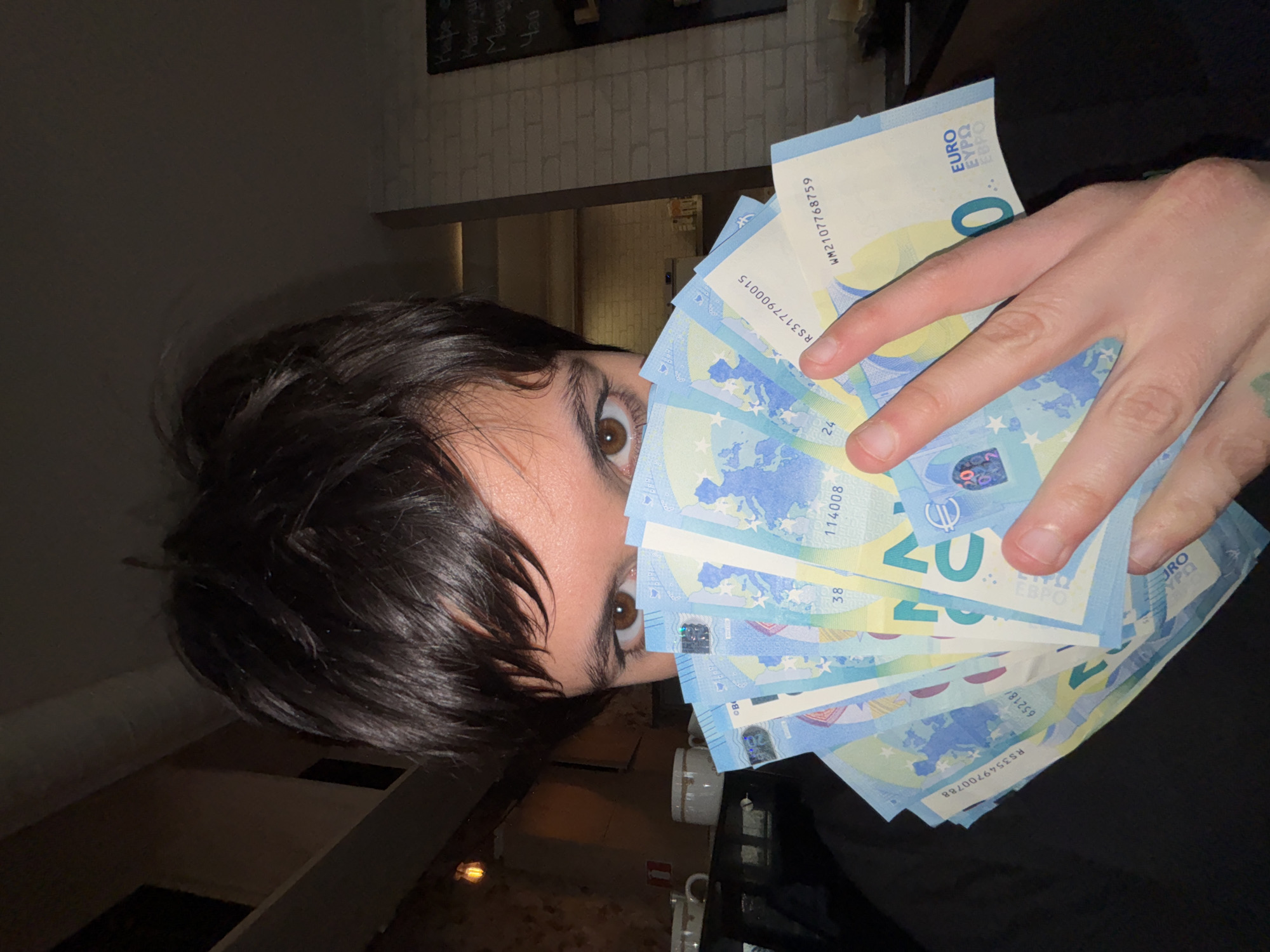 The image depicts a person holding a fan of 20 Euro banknotes in front of their face, partially obscuring it. Only one eye is fully visible, appearing brown. The person has short, dark hair. The banknotes are crisp and new, with serial numbers and holographic features visible.
The background suggests an interior space with a brick wall and a menu board listing "Кафе" (Coffee), "Марки" (Marks), "Manya" (possibly a transliteration of a name or drink), and "Чай" (Tea) in Bulgarian and Russian/Cyrillic script. Behind the person, there's a glimpse of a room or corridor with lighter-colored walls and furniture. Given the Bulgarian menu, a city location for Sofia, Bulgaria, is plausible. The scene appears to be indoors in a cafe.