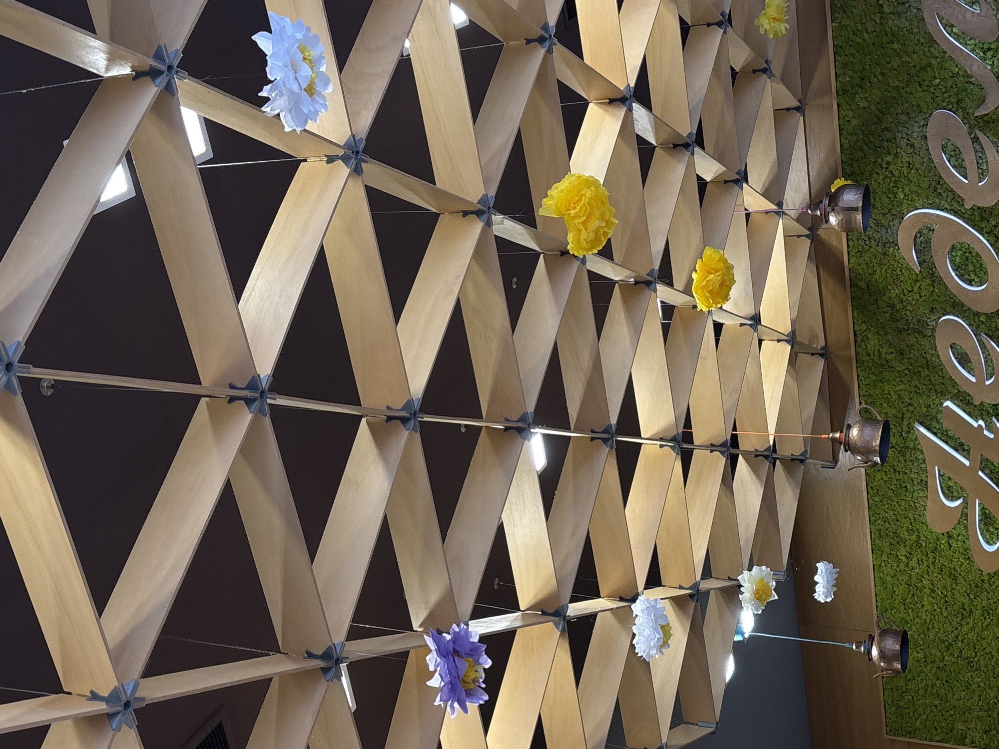 The image depicts an interior wall decoration. The wall features a geometric latticework made of light-colored wooden planks connected by gray fasteners. Paper flowers in various colors (white, yellow, purple) are interspersed throughout the lattice. A section of a green wall with white script or signage is visible on the right side. Metallic objects resembling small kettles are suspended from the latticework and are adjacent to the green wall section. The background behind the latticework is dark. The image does not contain people. The location in Varna, Bulgaria is not possible to determine from the image itself.