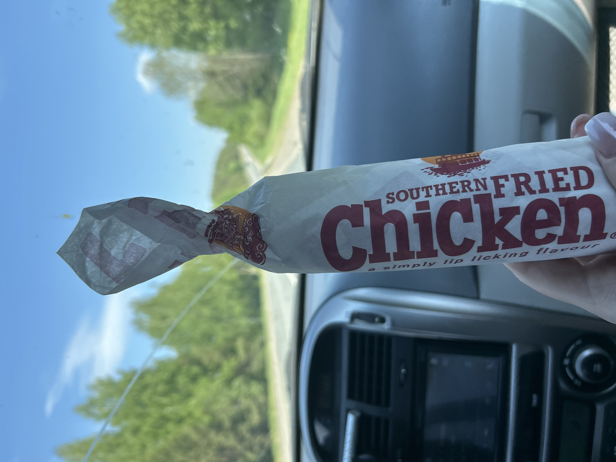 A person is holding a wrapped Southern Fried Chicken sandwich in a car. The interior of the car is visible, including the dashboard, vents, and a screen. Outside the car window, there is a view of trees and the sky. The location cannot be determined from the image.