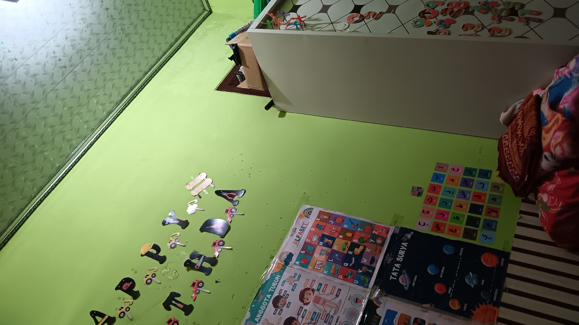 The image shows a child's bedroom in Sonobekel, Indonesia. The walls are painted a light green and are adorned with educational posters and alphabet decorations. One poster displays an alphabet chart with cartoon characters, while another features a solar system diagram with planets labeled in Indonesian. Arabic alphabet tiles are also visible on the wall. To the right, a large white wardrobe stands against the wall. The ceiling is a patterned white material. A bed with colorful bedding is partially visible in the foreground. The room appears to be well-lit, suggesting it's daytime. There are no people visible in the image.