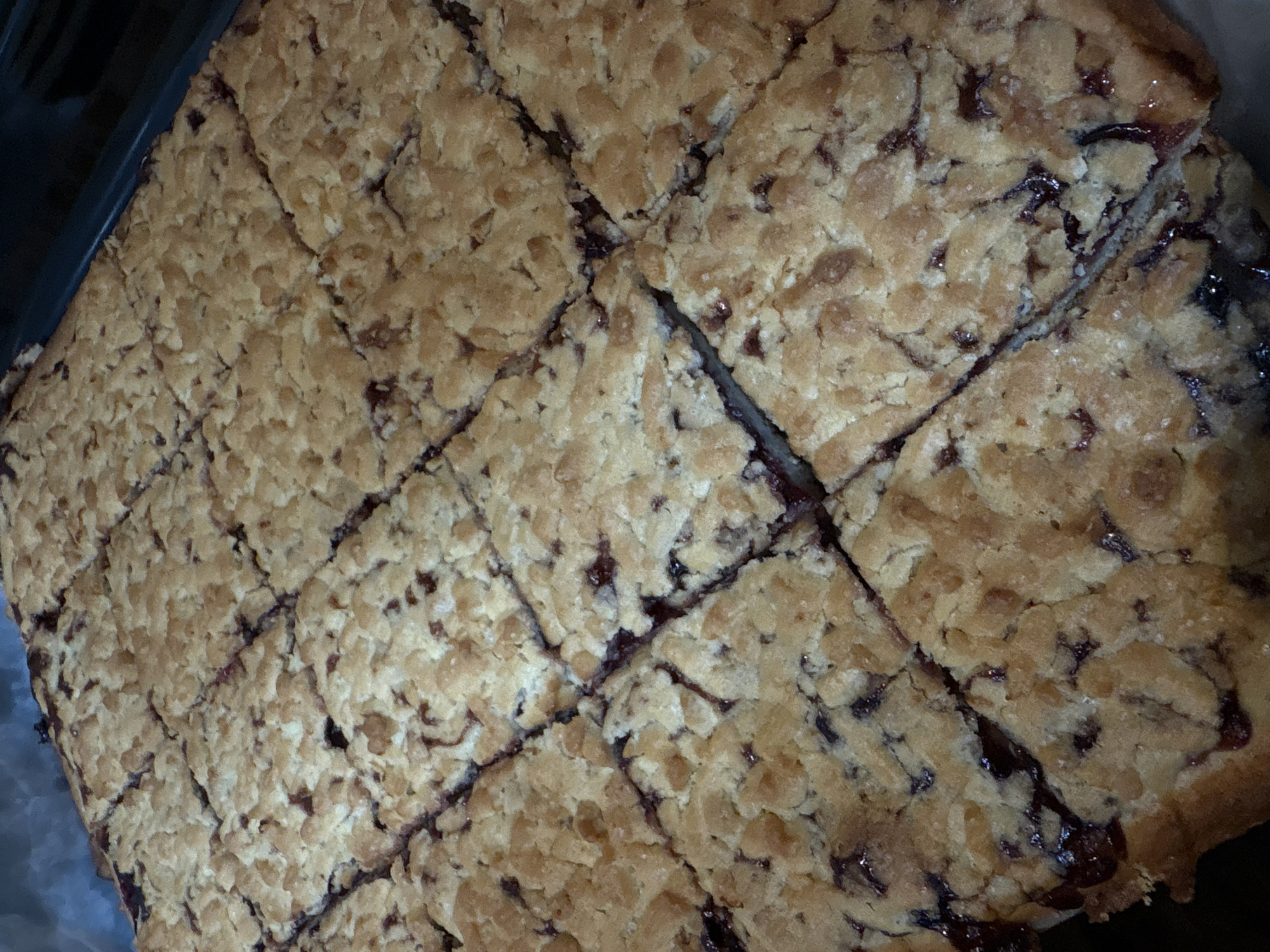 A close-up, overhead shot showcases a tray of freshly baked crumble bars, cut into squares. The bars have a golden-brown, crumbly topping interspersed with visible pieces of dark fruit filling, likely berries. The filling appears to be somewhat jam-like and peeks through the gaps in the crumble. The baked goods are arranged in a grid pattern, filling most of the frame, and appear to be on a dark surface, possibly a baking sheet or tray. The lighting is warm, highlighting the texture of the crumble. This image likely depicts a dessert or treat prepared in a kitchen setting in Vitsebsk, Belarus.