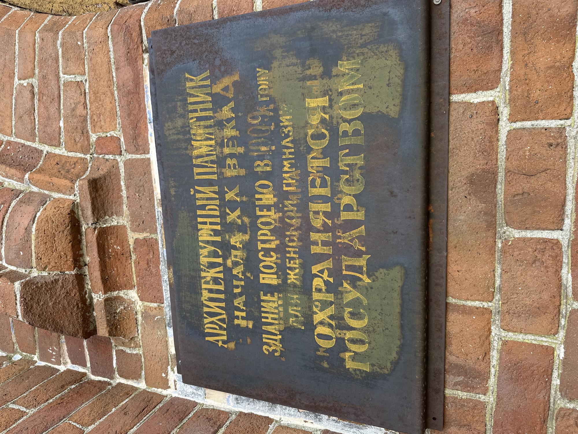 An exterior, eye-level, medium shot shows a weathered, dark metal plaque affixed to a red brick wall. The plaque is inscribed with yellow text in Russian, indicating it is an architectural monument. The text reads: "АРХИТЕКТУРНЫЙ ПАМЯТНИК НАЧАЛА ХХ ВЕКА ЗДАНИЕ ПОСТРОЕНО В 1909 ГОЛУ ДЛЯ ЖЕНСКОЙ ГИМНАЗИИ". Below this, a second line reads: "ОХРАНЯЕТСЯ ГОСУДАРСТВОМ". The plaque is slightly rusted and the text is faded in places. The brick wall shows signs of age and weathering. The image is taken in the city of Nogornyi, Russia.