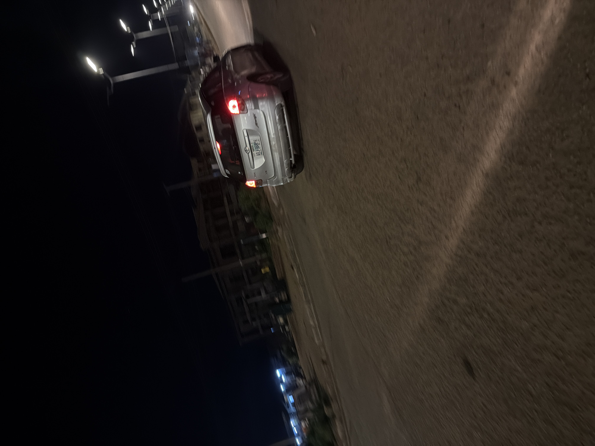 In Bukuru, Nigeria, at night, a silver Toyota hatchback is captured in motion on a paved road. The car's taillights are illuminated, indicating its movement. The background features several streetlights casting illumination on the scene, and the blurred shapes of buildings are visible. The road surface appears to be asphalt, with some dirt and debris. The image is slightly blurred due to the motion of the vehicle.