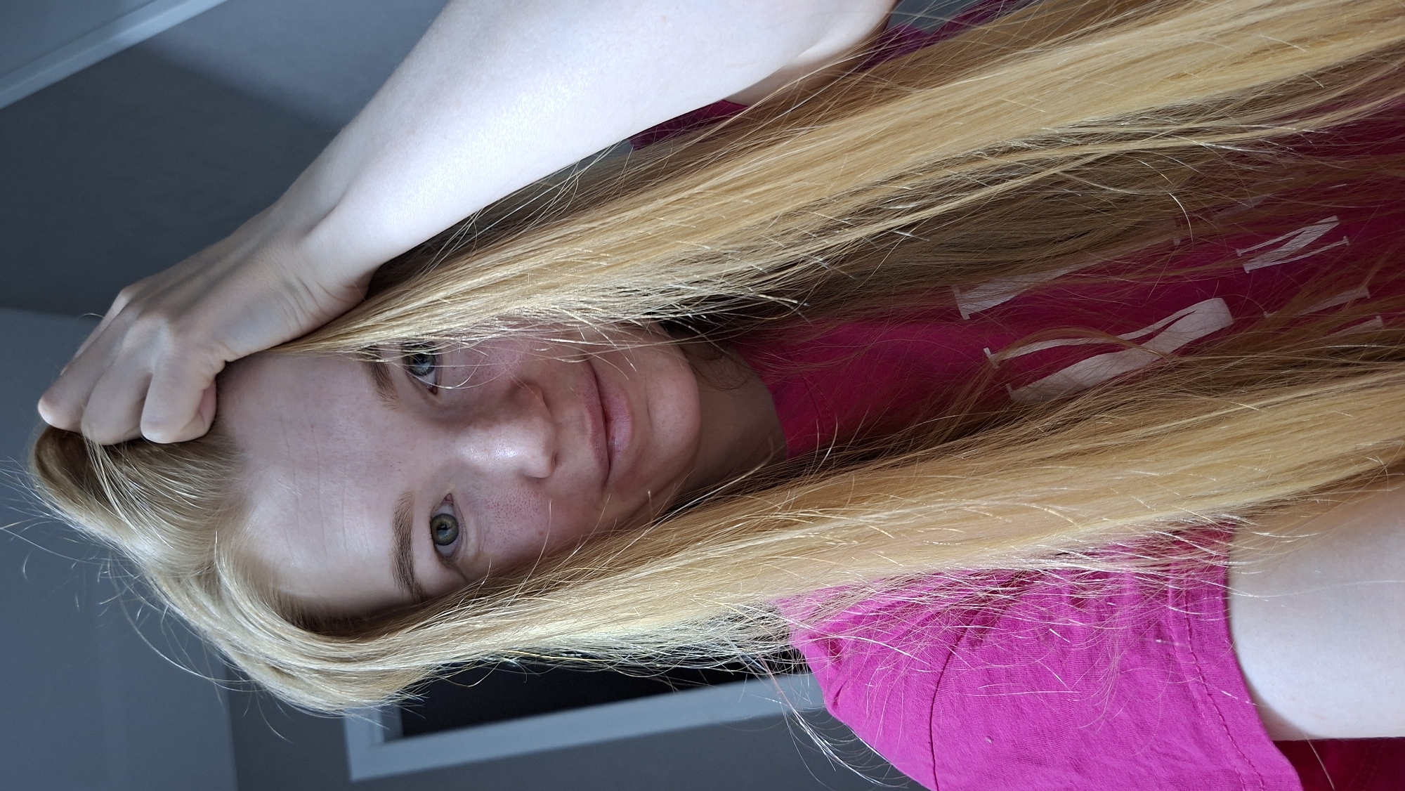 A person with long, blonde hair is positioned close to the camera, partially obscuring their face. They are wearing a bright pink t-shirt with white accents. Their left hand is raised near their forehead. The background is a neutral gray. There is no indication of the location being Chaikovsky, Russia.