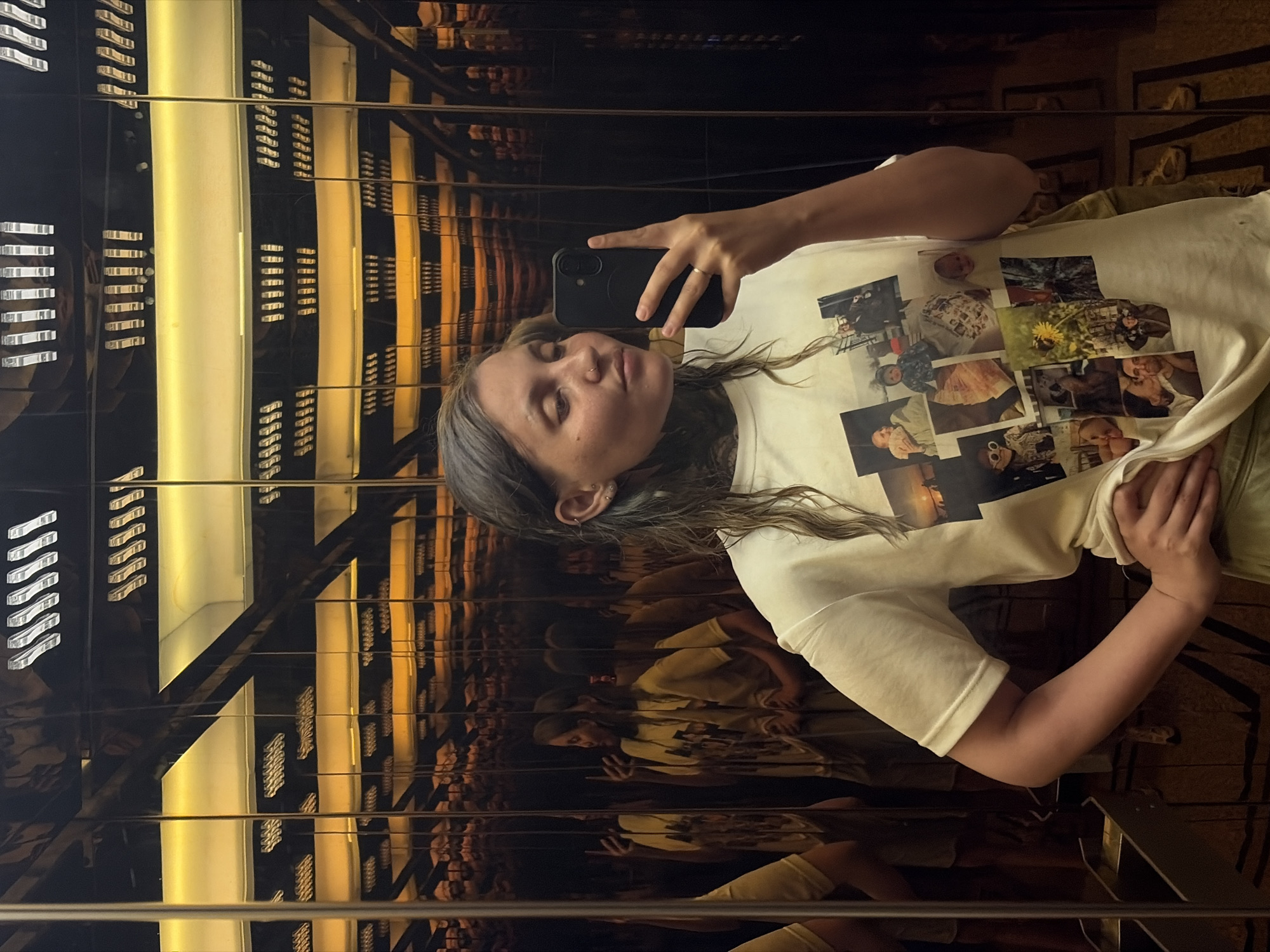 A young woman is depicted taking a selfie in a mirrored elevator or room. She is positioned diagonally, holding a dark-colored smartphone with a circular grip in her right hand. Her eyes are partially closed, and she has a nose piercing and a small stud earring, along with a metallic ring on her right ring finger. Her medium-length dark hair flows over her shoulders.
She is wearing a light-colored, possibly off-white or cream, short-sleeved t-shirt. The front of the t-shirt is decorated with a collage of printed photographs, predominantly featuring babies or young children in various poses and settings; one photo clearly shows a dandelion.
The setting is characterized by dark, highly reflective walls that create numerous, repeating reflections of the woman and the interior. Bright, warm yellowish light strips or panels illuminate the space, casting prominent horizontal reflections that contribute to a geometric and somewhat futuristic aesthetic. Metallic bars or railings are also visible, confirming an enclosed, artificial environment like an elevator. Faint reflections of another person can be seen deeper within the mirrored surfaces. The lighting is artificial and warm, creating an indoor ambiance. The image captures an everyday activity in an urban setting, specifically located in Đà Nẵng, Vietnam.