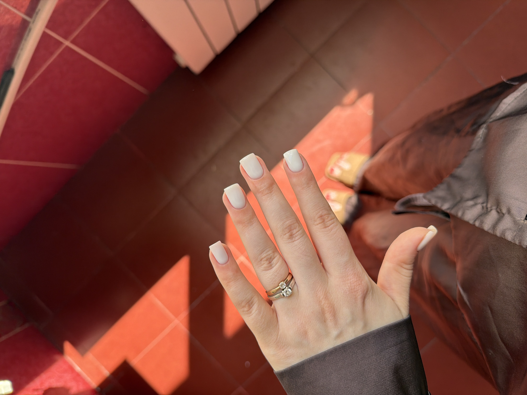 The image shows a close-up of a person's left hand, adorned with white-painted fingernails and two rings on the ring finger. The rings appear to be made of gold and silver, with at least one of them featuring a small diamond. The hand is resting against a dark-colored, textured fabric, possibly clothing.
The background consists of a tiled floor with a reddish-brown hue. Sunlight is casting strong shadows across the floor, suggesting it is daytime with bright illumination, possibly late afternoon due to the angle and length of the shadows. The setting appears to be indoors, given the tiled floor and the presence of what looks like furniture or shelving in the upper left corner, also illuminated by the sunlight. In the distance, a blurry object with yellow and white colors is visible, possibly shoes or slippers.
No text is visible in the image. The overall mood conveyed is casual and relaxed, with a focus on personal adornment. The location context of Kyiv, Ukraine, does not provide specific visual cues within the image itself.