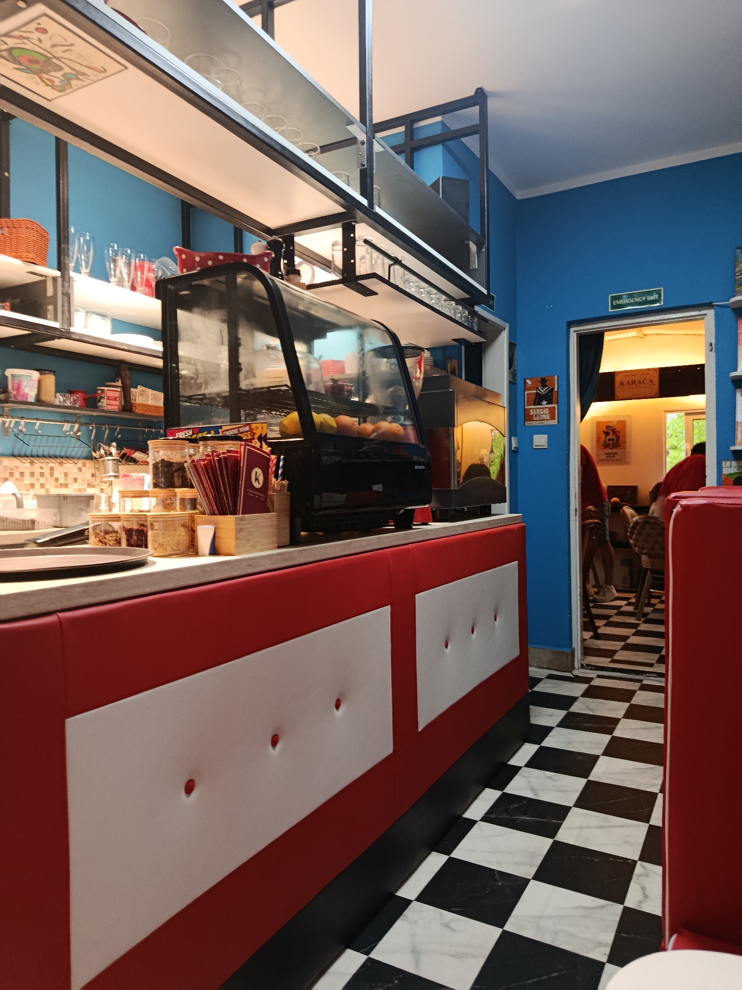 This media file captures the interior of a vibrant cafe or diner, likely located in Herceg Novi, Montenegro, characterized by a distinct retro aesthetic. The scene is viewed from inside, looking towards a service counter and an adjoining room.
In the foreground, a striking black and white checkered tile floor dominates, extending throughout the visible areas. Red and white padded booth seating with white rectangular panels and small circular indentations flanks the view.
The mid-ground features a well-equipped service counter with a light-colored top. A glass display case showcases items like pastries or fruits. Behind the counter, against a bright blue wall, shelves are neatly arranged with various supplies: clear jars containing dry goods, stacks of red-bound menus or notepads, straws, and a coffee machine. Overhead, more open shelving holds glassware and other cafe necessities.
To the right, an open doorway leads into another room, which continues the theme with red seating and the checkered floor. Patrons in red attire are partially visible, seated at tables with wooden chairs. On the blue wall adjacent to the doorway, two framed posters are displayed: one featuring a cowboy figure and the name "SERGIO LEONE," and another above it with the text "KARAÇA." Directly above the doorway, a green sign indicates "Emergency Exit." On the counter, visible text includes "FRESH" on a box and a "K" logo on a container of straws or menus, with partially visible text "CHOOSE YOUR...".
The lighting is consistent with an indoor setting during daytime. The overall ambiance is cheerful, clean, and inviting, suggesting a casual dining or cafe experience.
