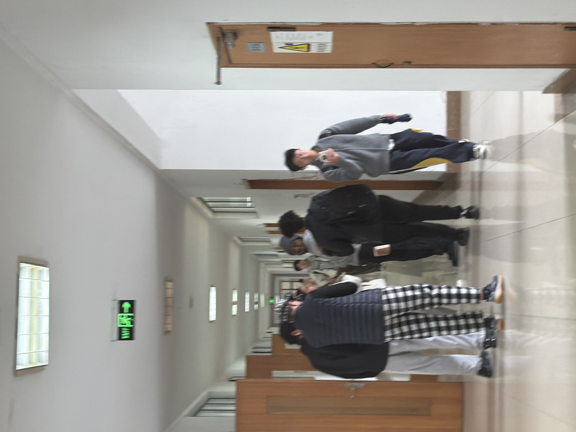 Here's a detailed description of the image:
**Location:** Appears to be an interior corridor in a building, possibly in Jiangning District, China. This assessment is based solely on visual analysis and the absence of information confirming a specific location.
**Scene:** The image depicts a long, straight corridor with tiled flooring. The walls are white, and there are evenly spaced windows or light fixtures along the left side. There are benches along the right wall.
**People:** There are multiple people walking down the corridor, generally in the same direction.
* The person closest to the camera is wearing a black jacket, a grey and white checkered skirt, and grey pants.
* The second person is wearing a black jacket and pants.
* The third person is wearing a grey hoodie and navy pants.
**Objects:**
* Windows/Light Fixtures: Present on the left side of the corridor.
* Benches: Lined along the right side of the corridor.
* Door: At the far end of the corridor, there is a brown door with a warning sign.
* Exit Sign: A green exit sign is visible on the left wall.
**Actions/Interactions:** The people are walking down the corridor.