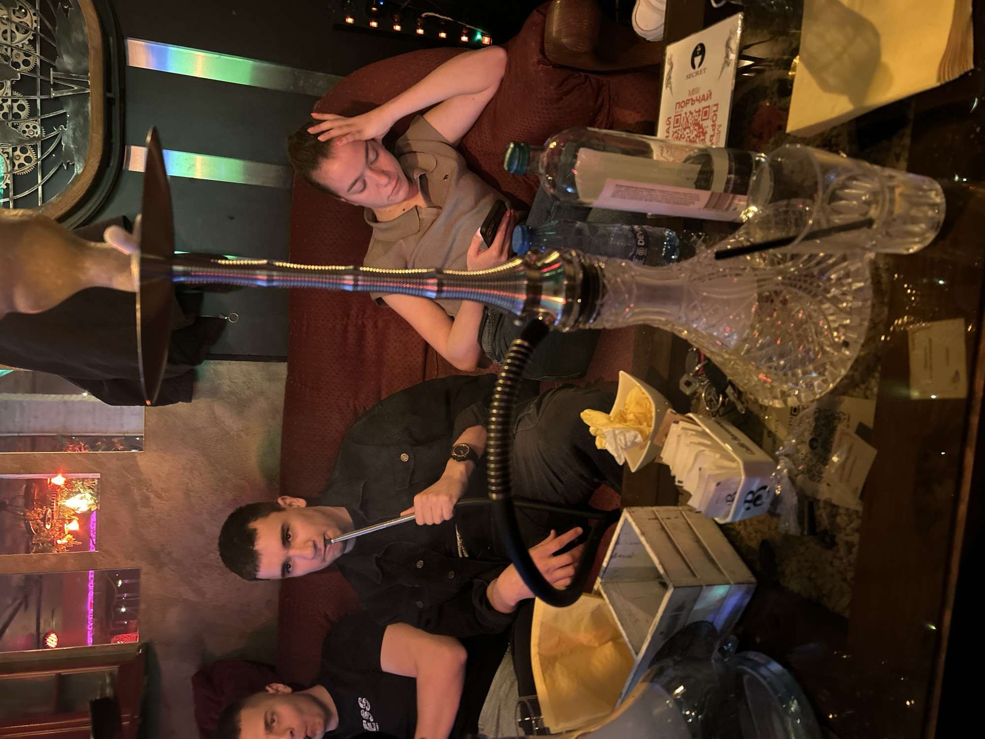 Here is a description of the image:
**Scene:** An indoor scene, likely a lounge or a bar in Pleven, Bulgaria, featuring a group of people relaxing and using a hookah. The image is rotated counter-clockwise by approximately 90 degrees.
**People:**
* A woman is reclined on a burgundy velvet couch, possibly in a state of relaxation or introspection. She's wearing a neutral-colored collared shirt and jeans. She is holding a smartphone in her hand.
* A man is seated and holding the hookah hose, about to take a drag. He is wearing a dark jacket.
* Another person is partially visible near the bottom of the frame wearing a black t-shirt.
**Objects:**
* A hookah is centrally placed in the frame, consisting of the base (a decorative crystal glass), the stem, the hose, and the bowl.
* The hookah is placed on a dark glass table.
* Bottles of water and other containers are present on the table. A menu card from "Secret Room" is also on the table.
* A wooden box with a yellow plastic bag inside sits near the bottom of the frame.
* Sugar packets and other miscellaneous items are scattered around the table.
* A decorative wall piece is visible in the background, featuring gears and metallic accents.
* Mirrored panels are seen on the wall, reflecting neon and purple lighting.
**Actions/Interactions:**
* People are smoking hookah.
* The woman is looking at her phone.
* The subjects appear to be in a relaxed environment.
**Environment:**
* Low lighting with neon lighting.
* The décor and furniture suggest a modern or industrial-chic style.
* The lounge/bar setting hints at an entertainment or social gathering place.
* Overall, the scene suggests a gathering of friends in a relaxed atmosphere enjoying a hookah and drinks in a stylish venue.